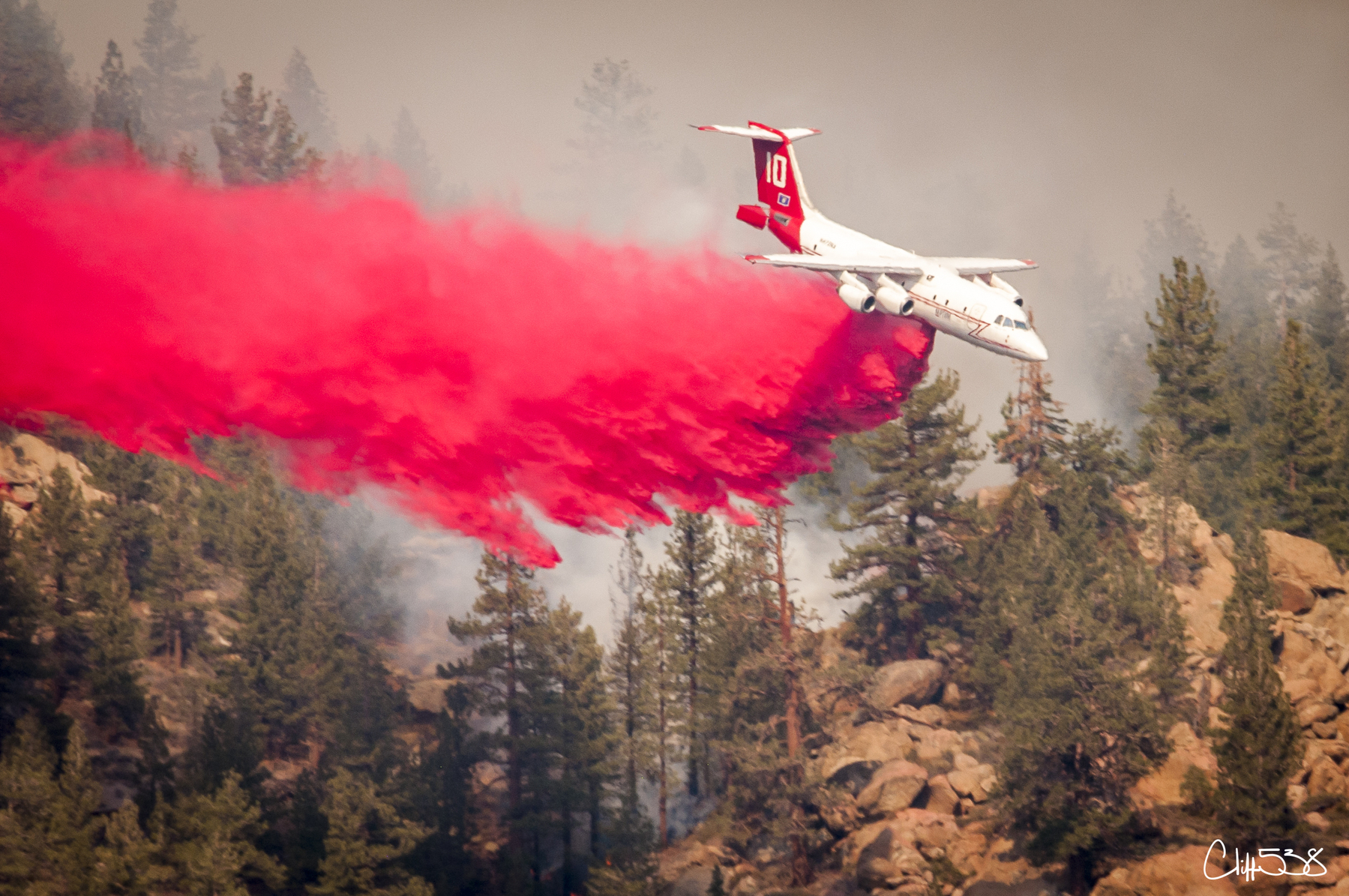 Auto-generated description: A firefighting plane releases a bright red fire retardant over a forested area with smoke rising from the trees.