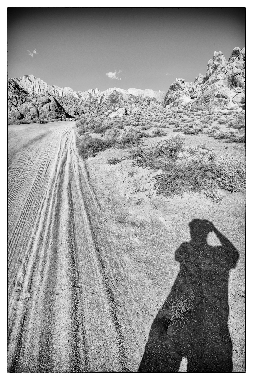 Auto-generated description: A black and white scene featuring a shadow of a person taking a photo in a desert landscape with mountains and a dirt road.