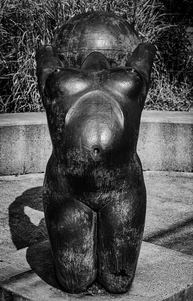 Auto-generated description: A bronze sculpture of a kneeling figure holding a large spherical object above its head is displayed outdoors with grass and a concrete bench in the background.