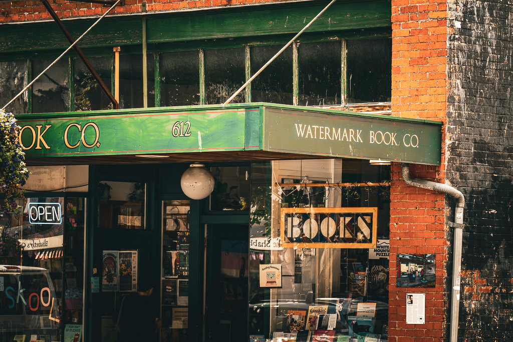 Auto-generated description: A quaint bookstore named Watermark Book Co. with a green awning and a hanging BOOKS sign is located on a brick building.