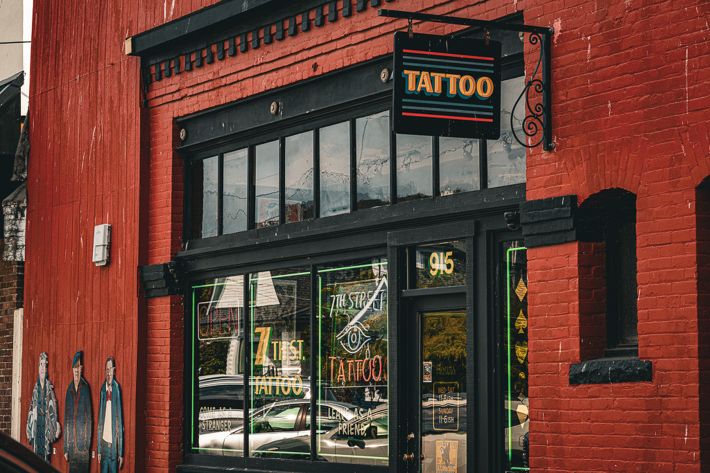 Auto-generated description: A tattoo shop with a black and orange sign is located in a red brick building, featuring large windows and artwork on the adjacent wall.