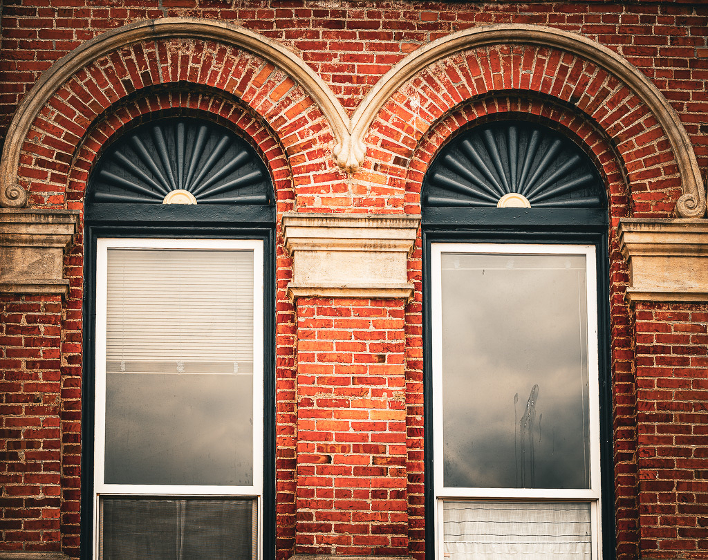 Auto-generated description: Two arched windows with decorative fan-shaped transoms are set in a red brick wall.