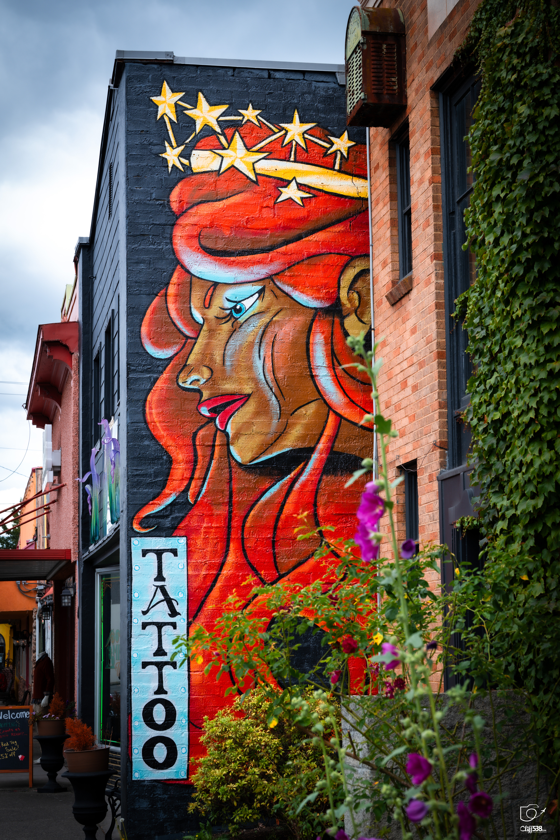 Auto-generated description: A vibrant mural of a woman with flowing red hair and a crown of stars adorns the side of a tattoo shop building.
