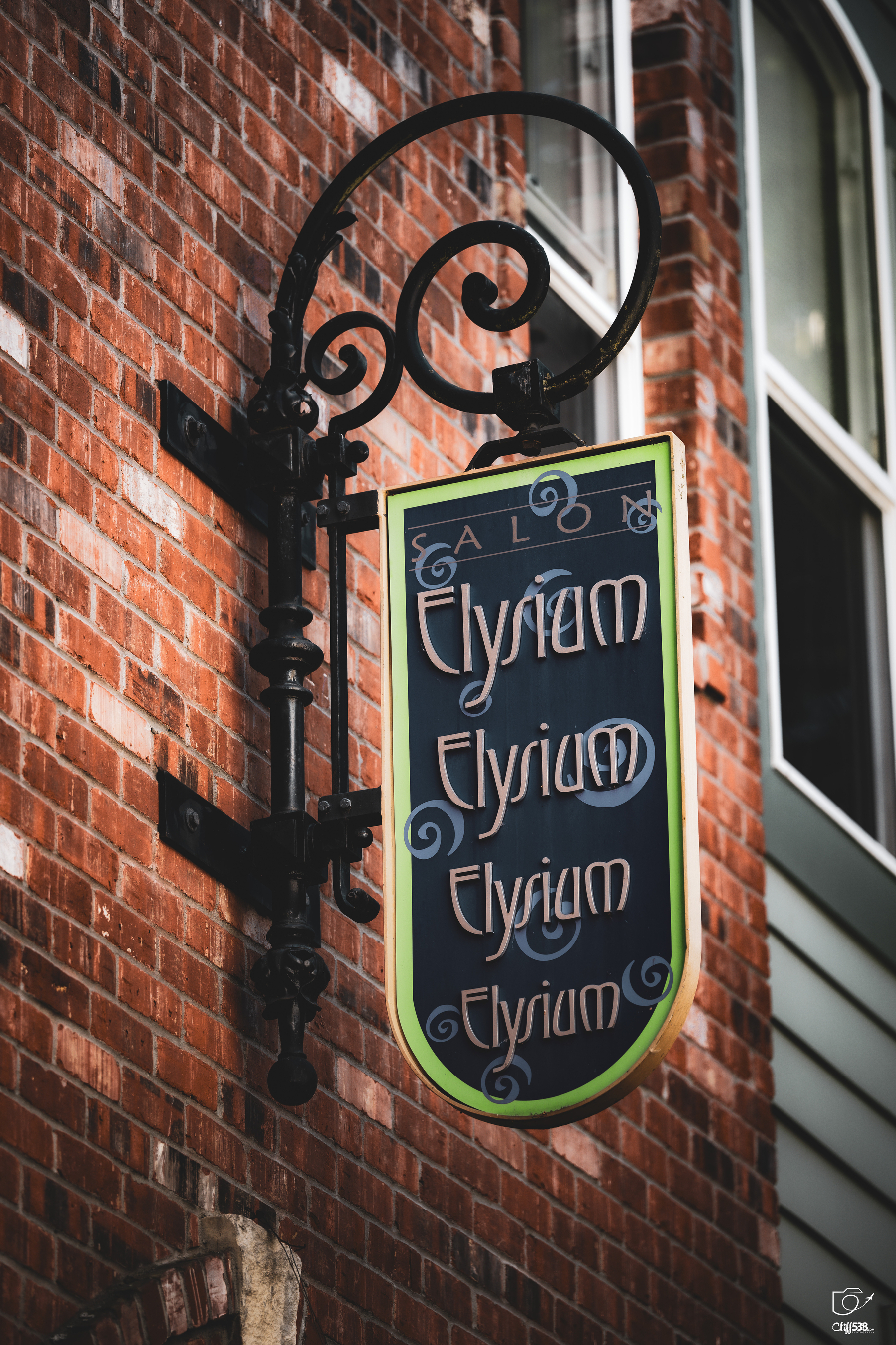 Auto-generated description: A decorative hanging sign with the word Elysium repeated multiple times is mounted on a brick wall next to a window.