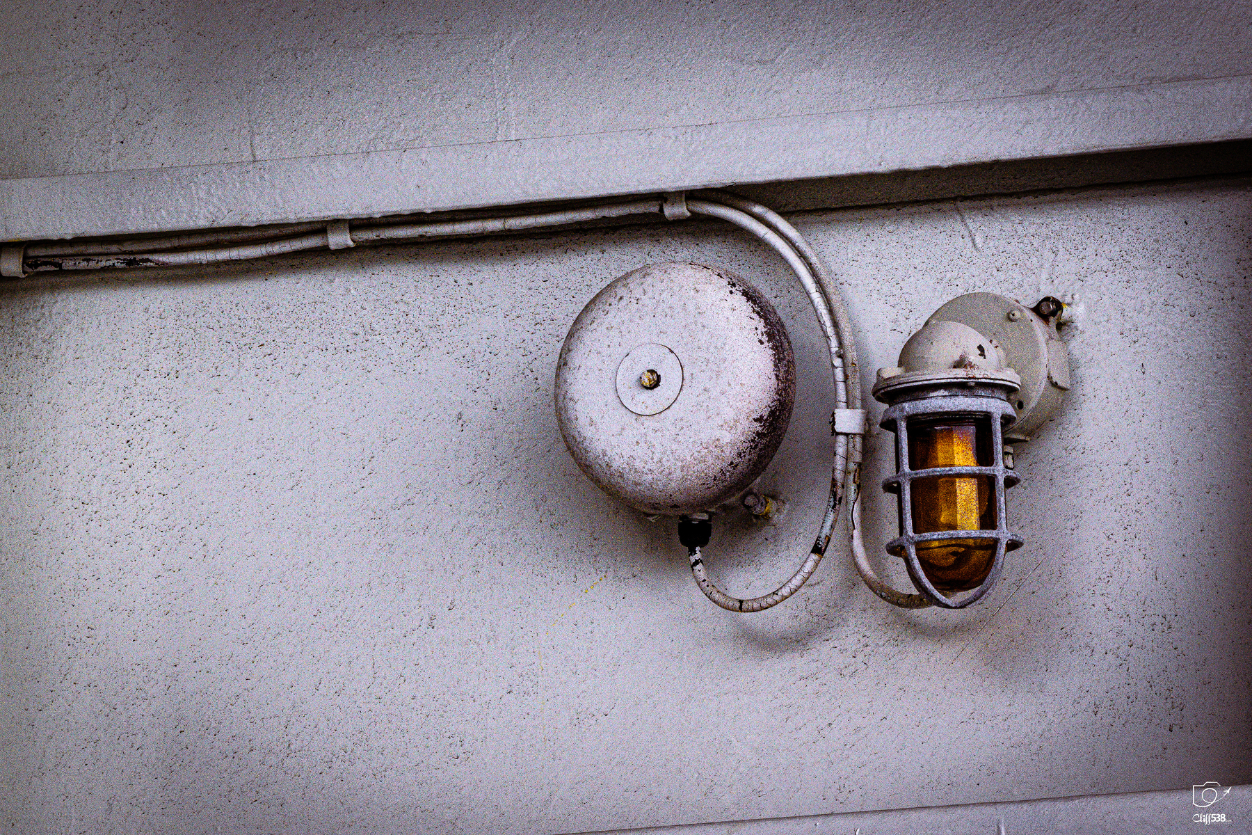 Auto-generated description: A rusty old fire alarm bell and a protective cage-covered light fixture are mounted on a worn white wall.