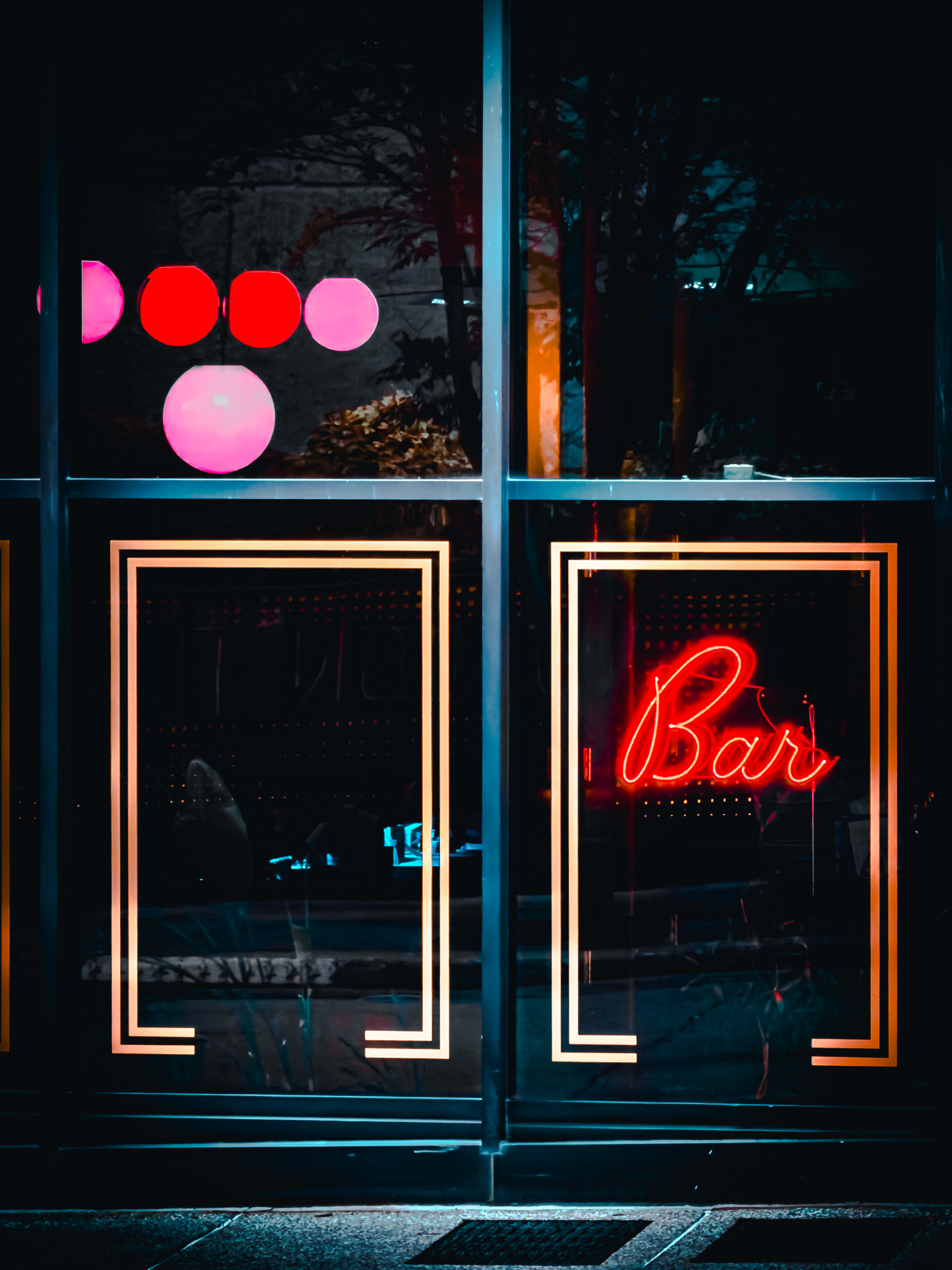 Auto-generated description: A glass door with illuminated rectangular borders features a bright red neon sign that reads Bar, set against a dark background with reflections of colorful circular lights.