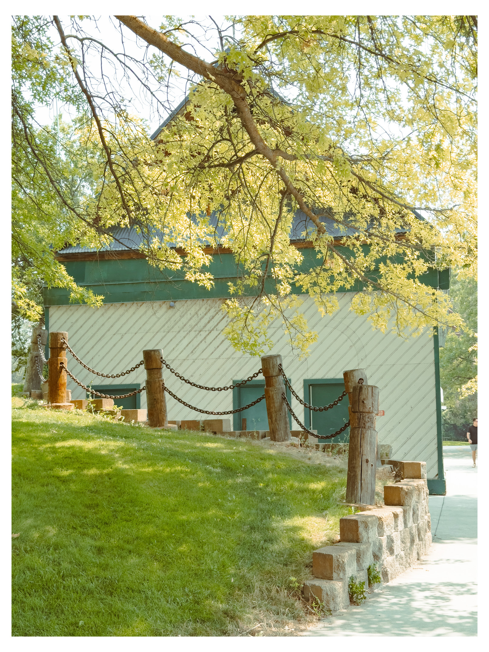 Auto-generated description: A sloped grassy area with a chain-linked wooden post fence leads up to a green and white building partially shaded by leafy tree branches.