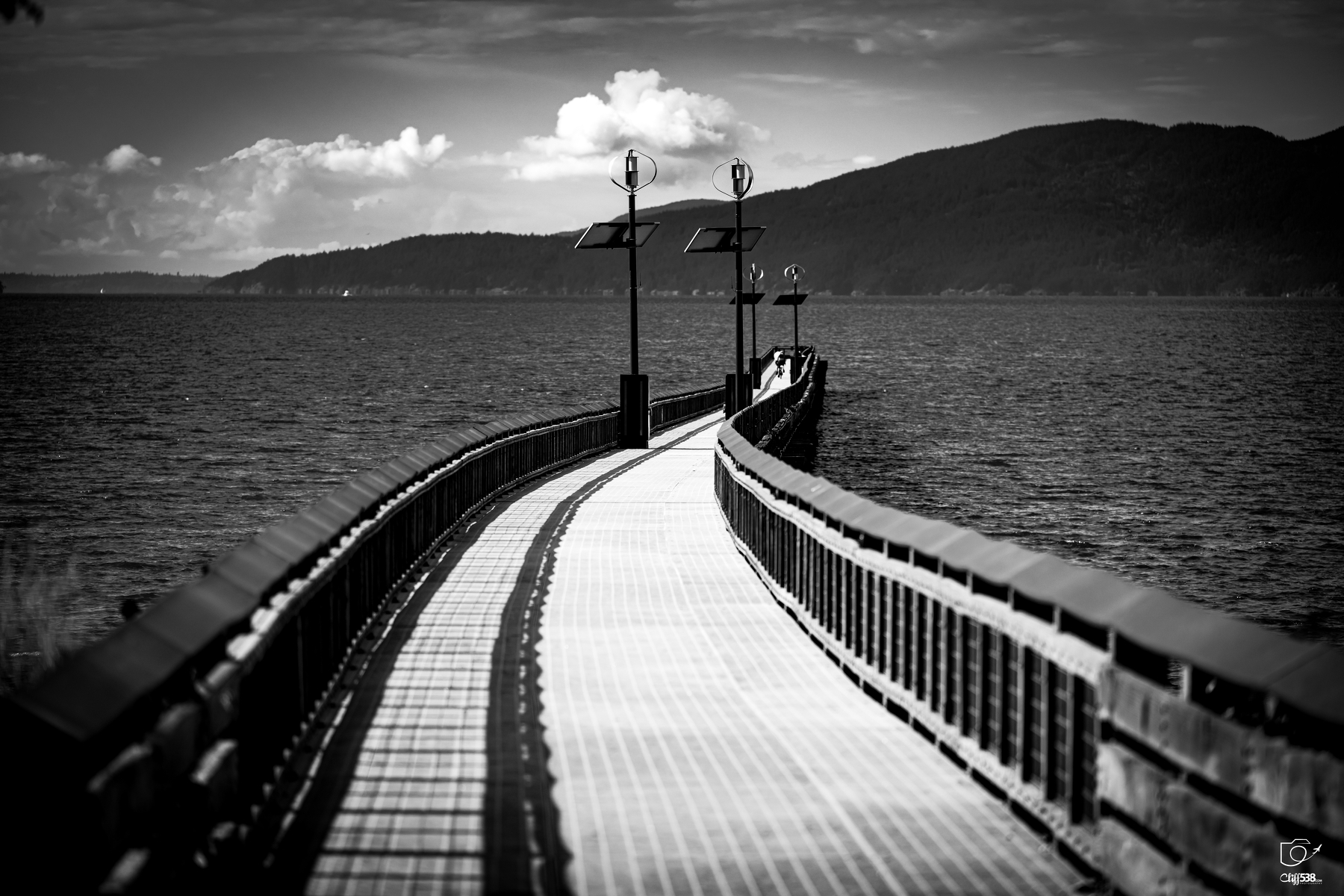 Auto-generated description: A curved wooden pier extends over a body of water towards a distant mountainous shoreline under a partly cloudy sky.