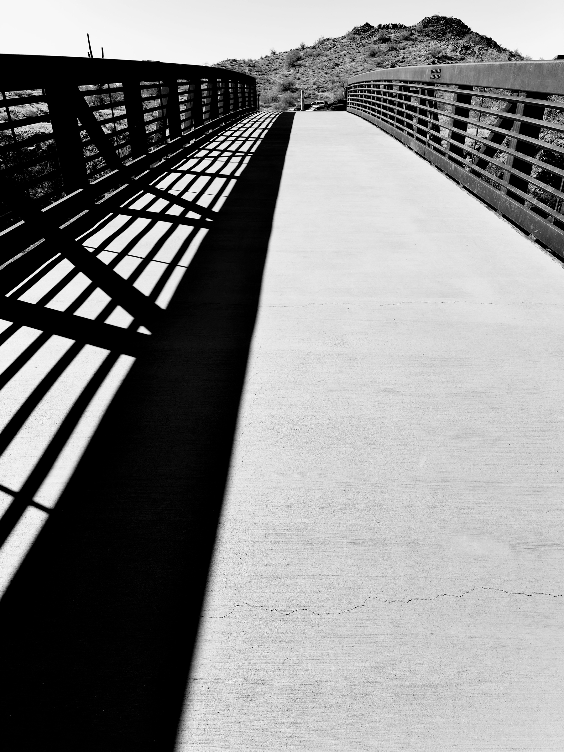 Auto-generated description: A bridge casts long, geometric shadows over its walkway, leading towards distant hills under a clear sky.