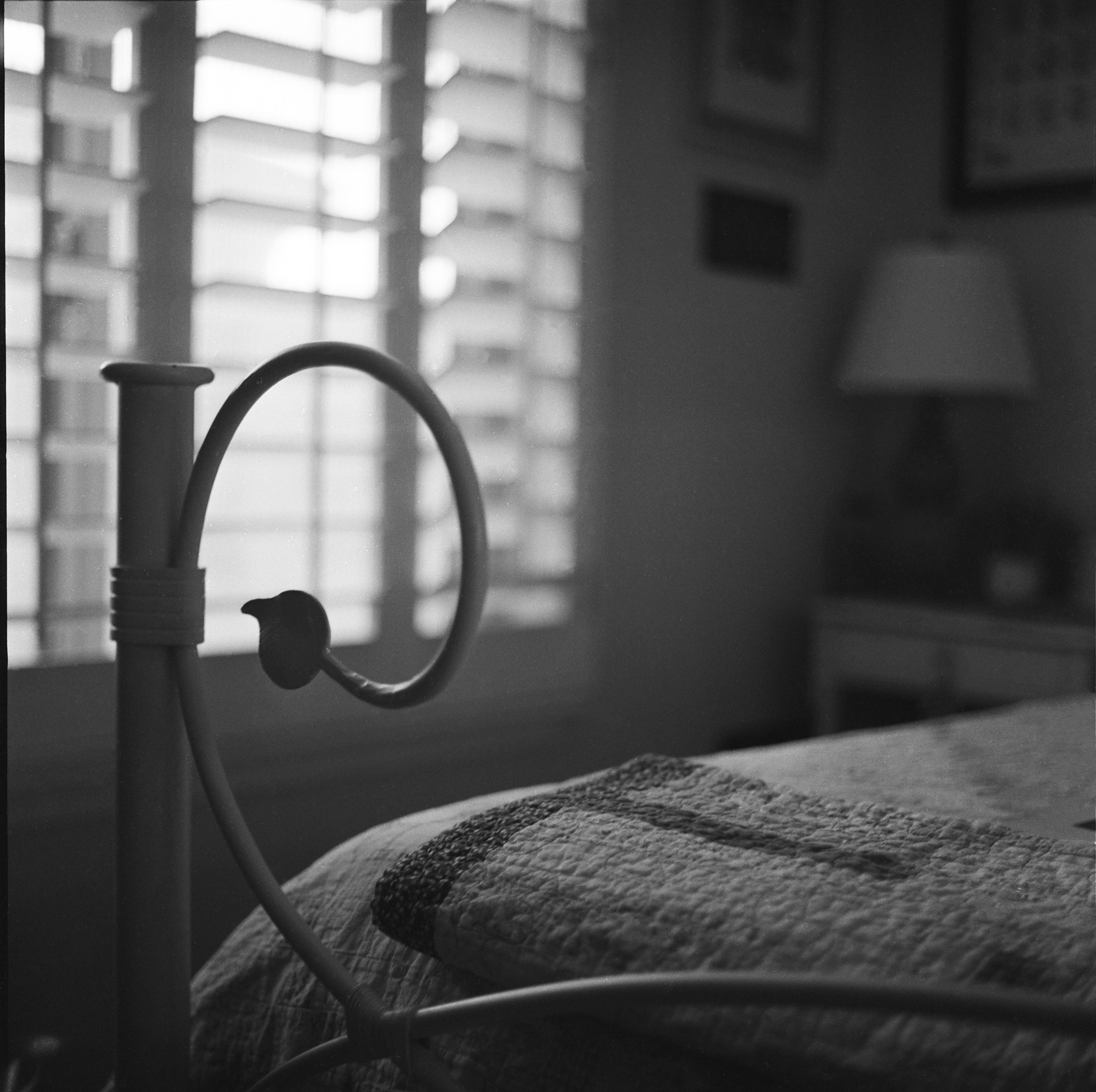 Auto-generated description: A close-up of a cozy bedroom corner features an iron bed frame with a bird-shaped detail and a quilted blanket, softly lit by natural light from a window with blinds.