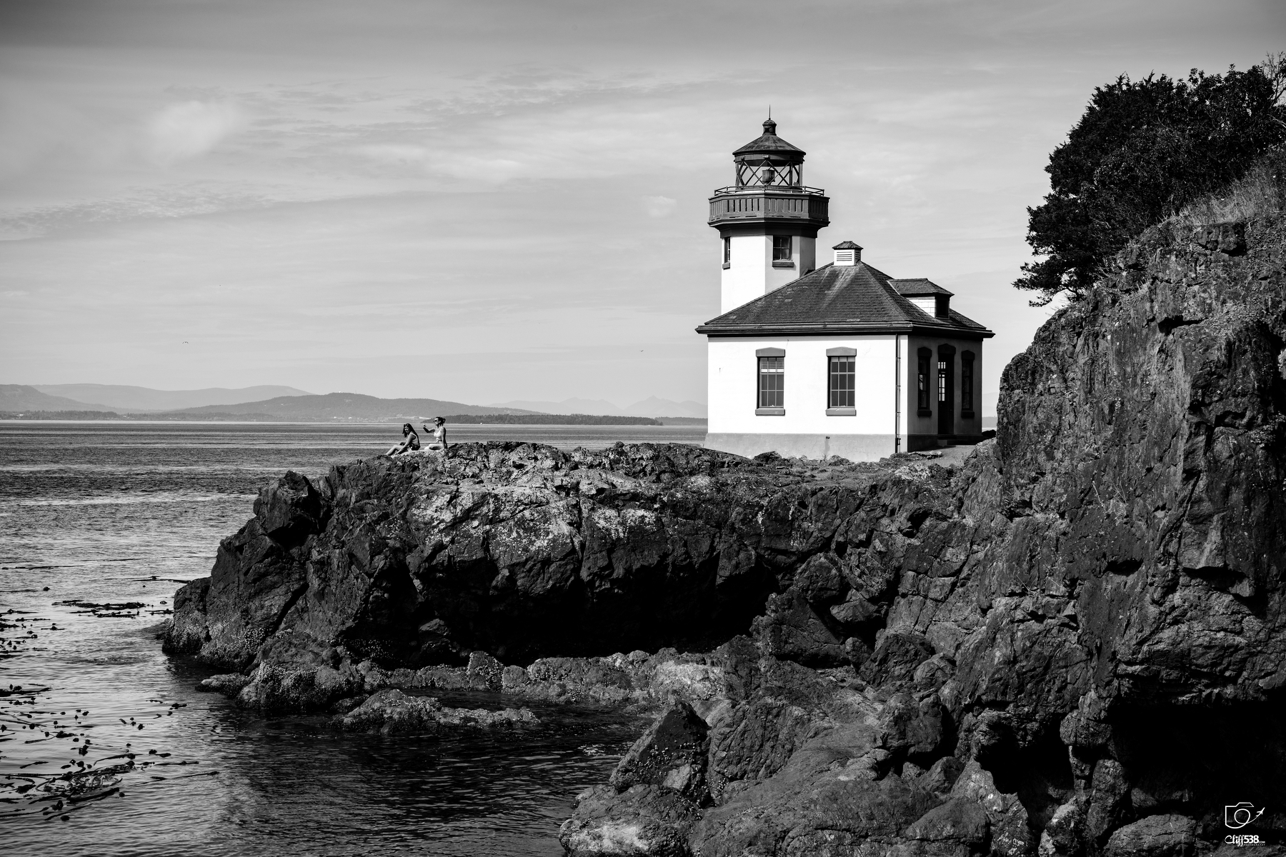 Auto-generated description: A lighthouse stands on a rocky cliff overlooking a calm body of water under a partly cloudy sky.