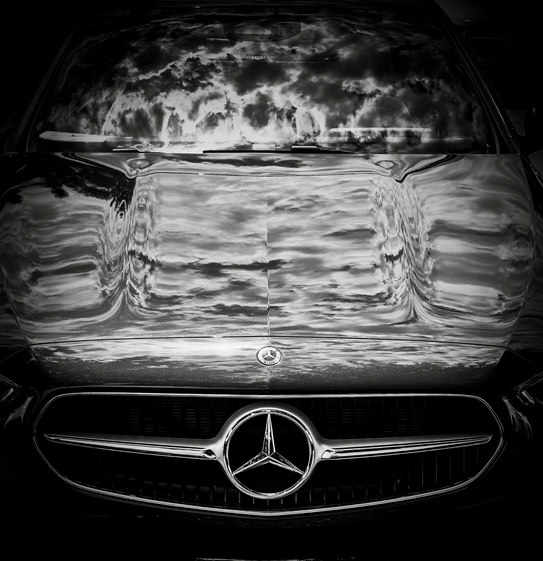 Auto-generated description: A close-up of a Mercedes-Benz car hood and grille reflects a dramatic, cloudy sky.