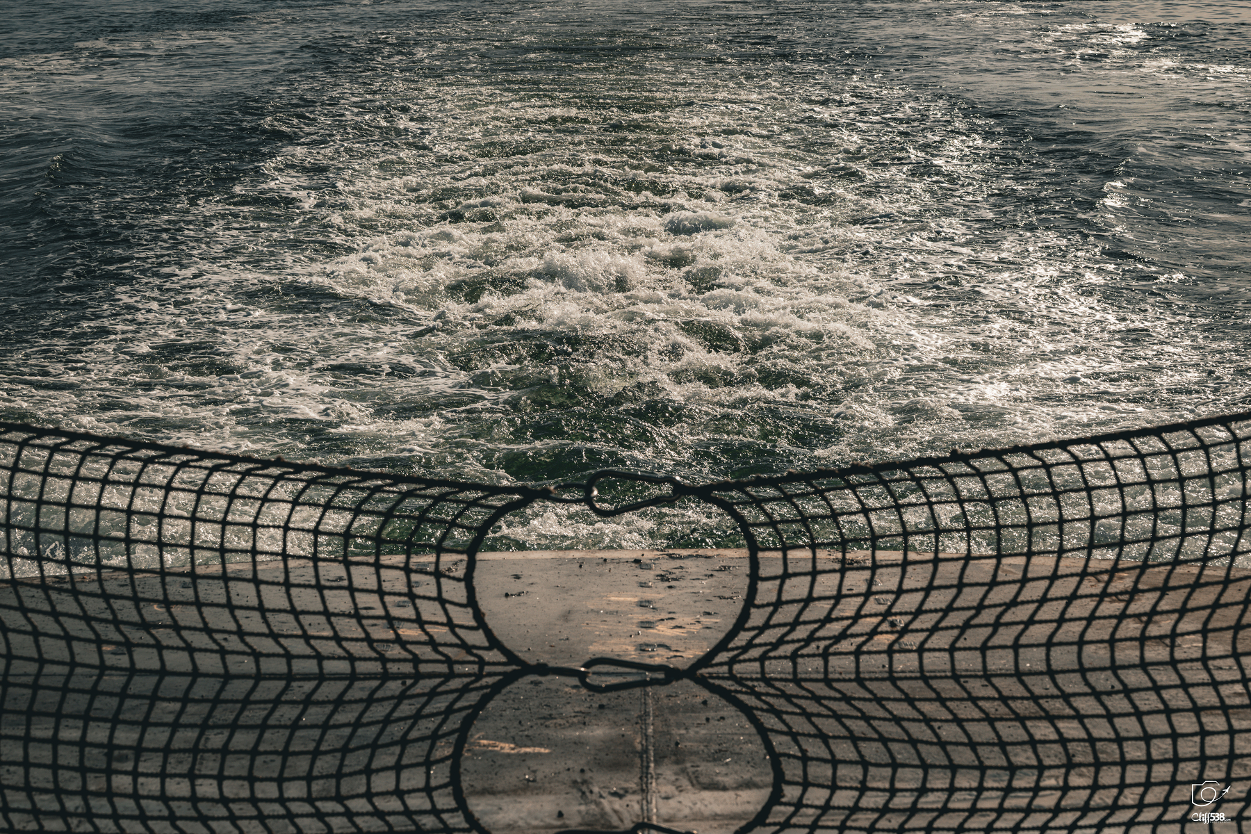 Auto-generated description: A view of turbulent water behind a boat, partially obscured by a safety net attached to the rear railing.