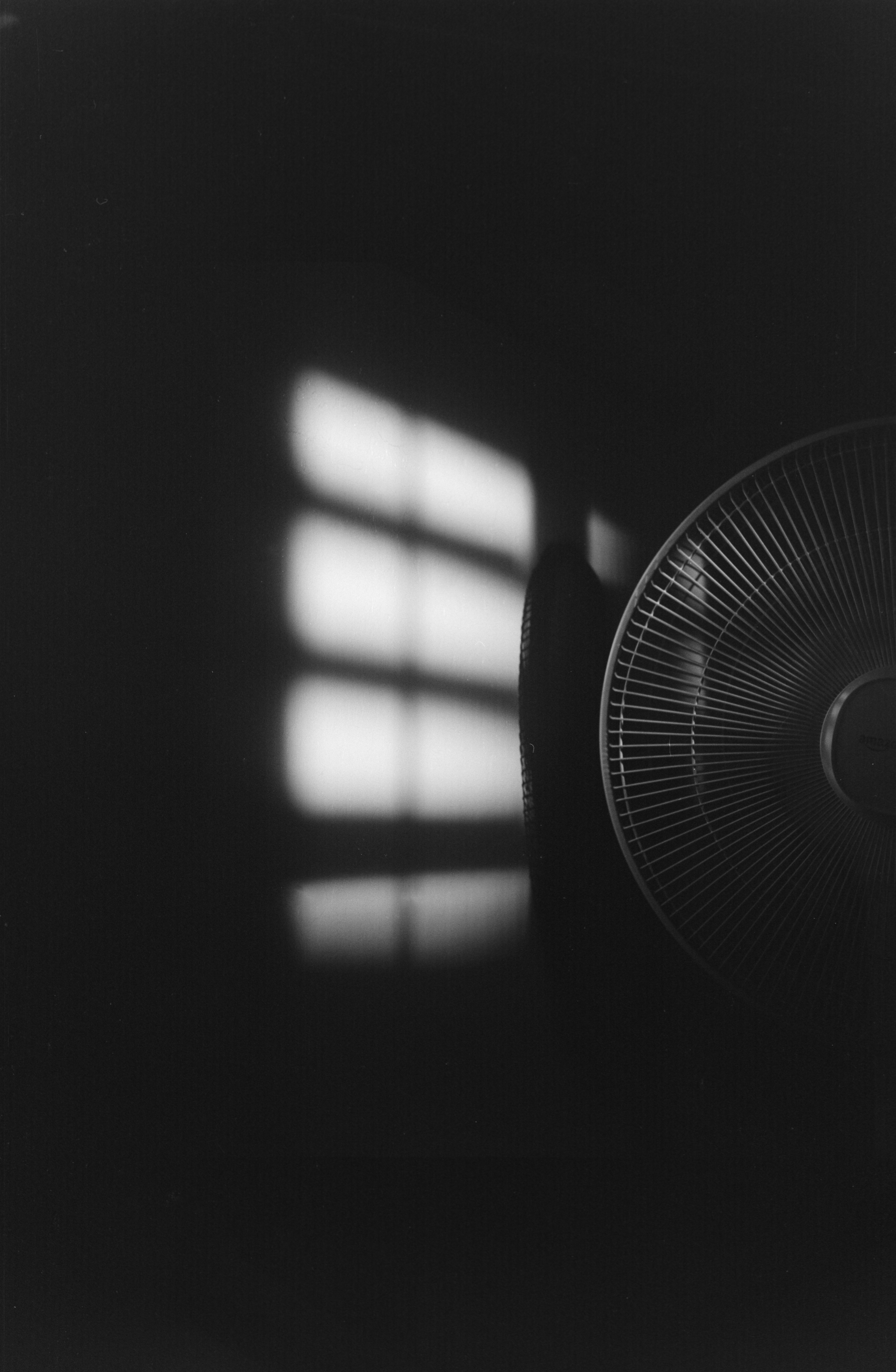 Auto-generated description: Soft light filters through a window and casts a grid-like pattern on a dark wall beside a fan.