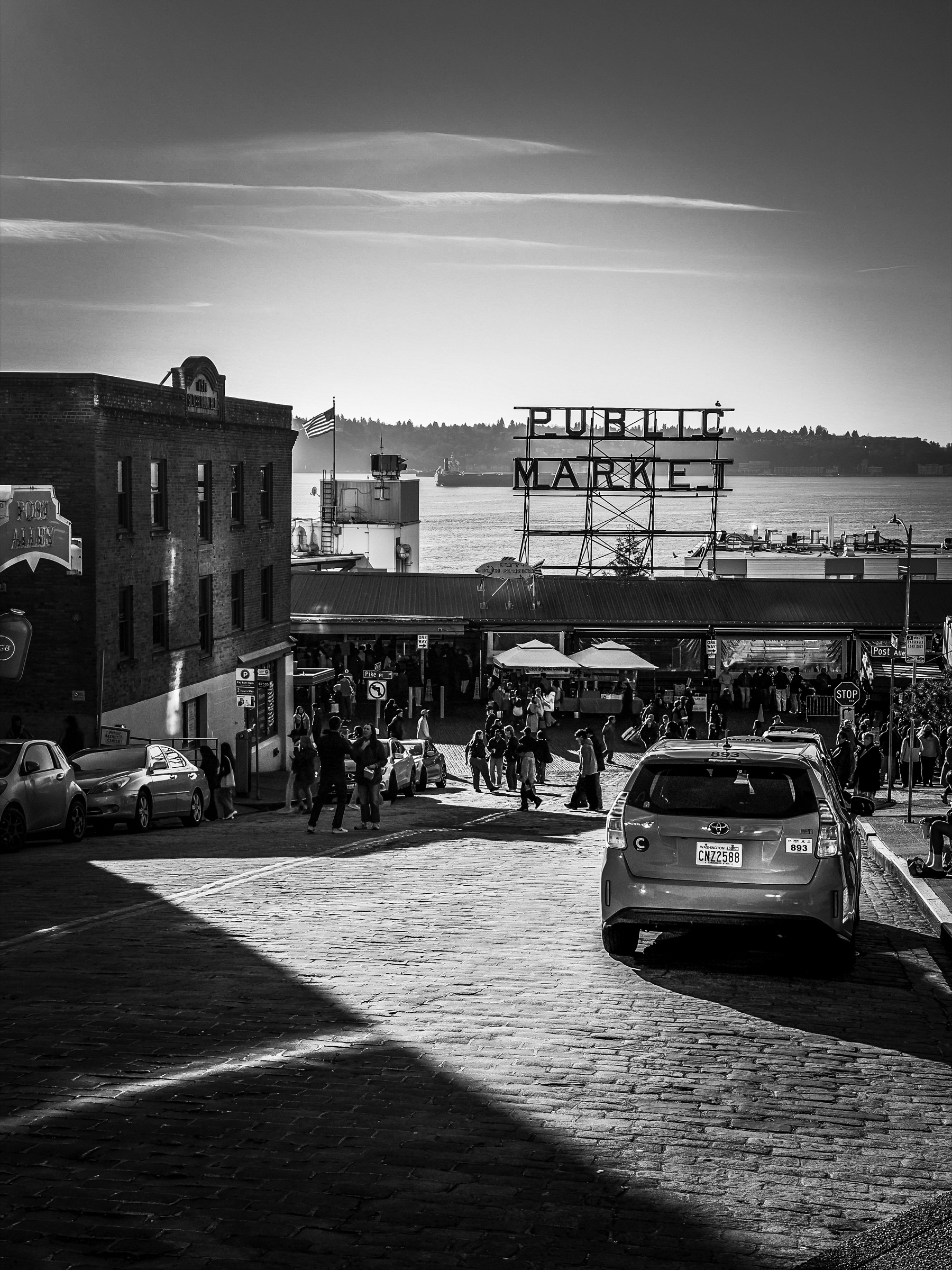 Auto-generated description: A bustling street scene in black and white captures a historic public market with people walking and cars parked, overlooking a waterfront.