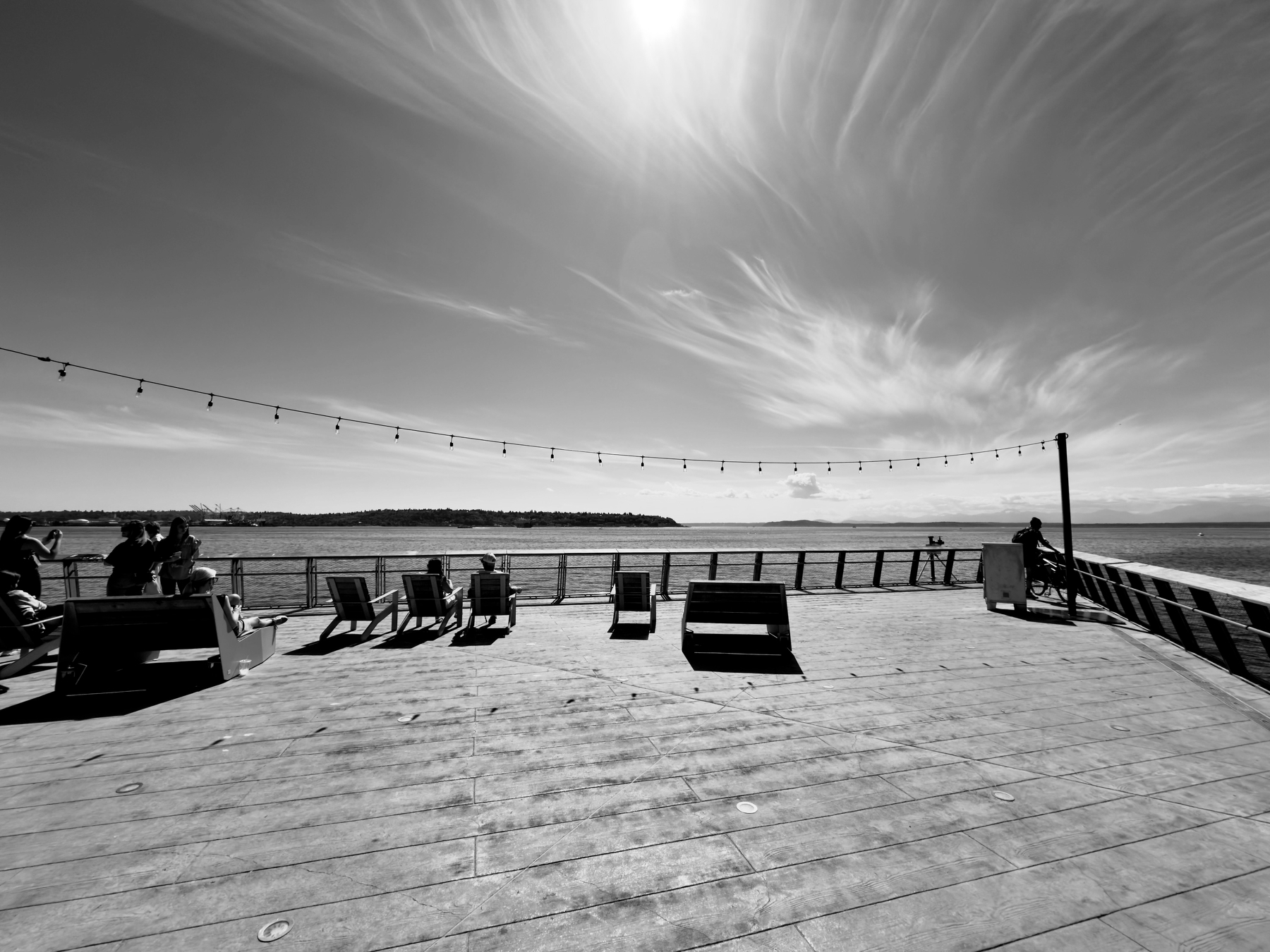 Auto-generated description: A waterfront boardwalk features people relaxing on benches under a string of lights, with a picturesque view of water and clouds in the sky.