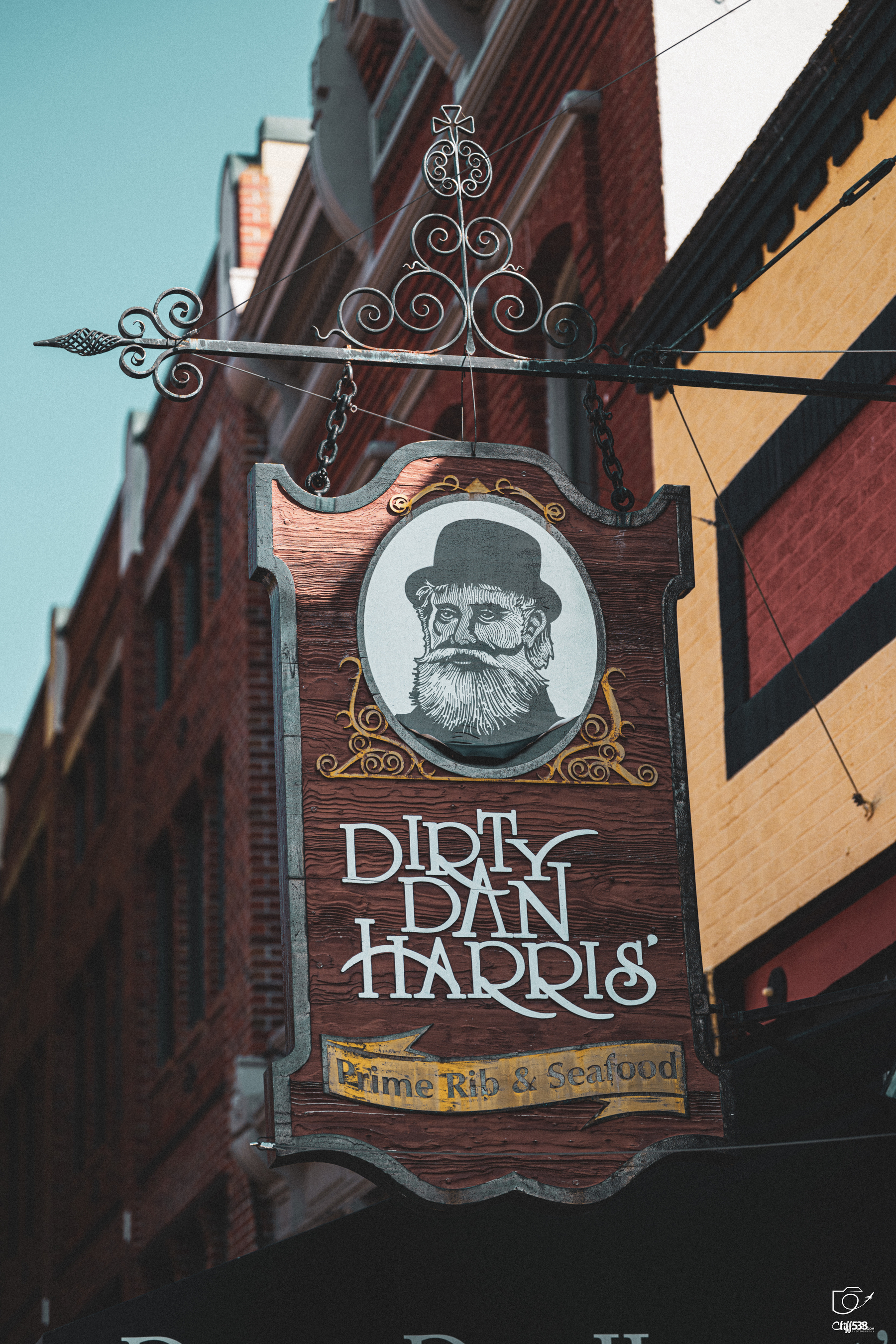 Auto-generated description: A wooden hanging sign for Dirty Dan Harris' featuring an illustrated bearded man in a top hat advertises prime rib and seafood outside a brick building.