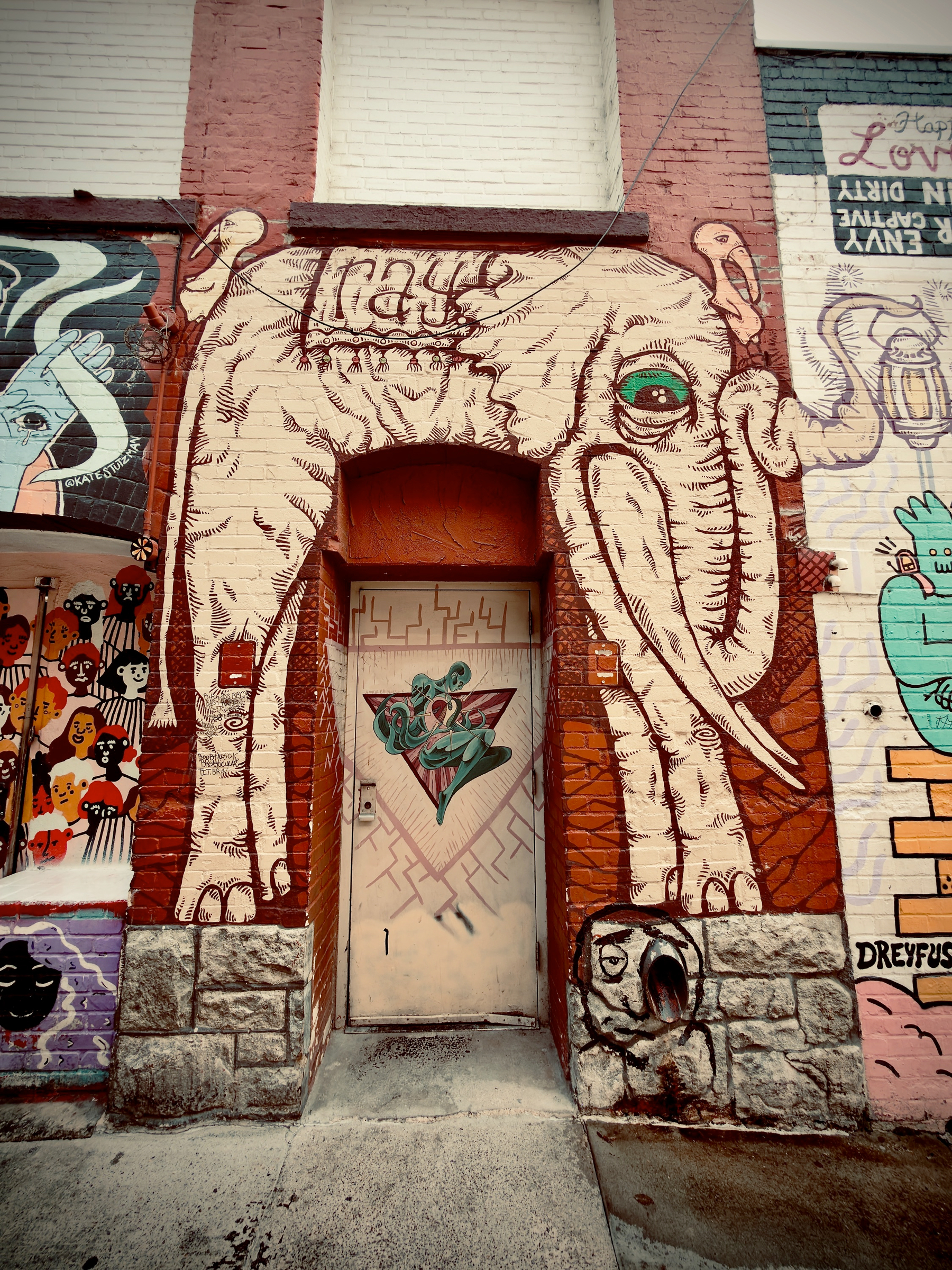 Auto-generated description: A large elephant mural painted around a door on a brick wall features detailed line work and an expressive eye, surrounded by other colorful street art.