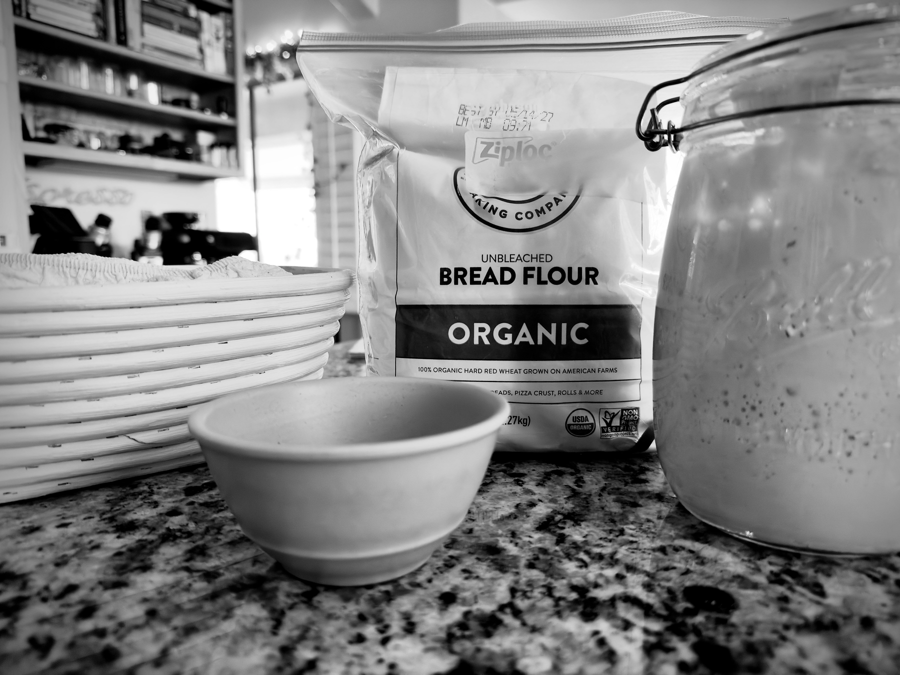 Auto-generated description: A bag of organic unbleached bread flour sits on a countertop next to a small bowl and a jar, with a basket partially visible in the background.