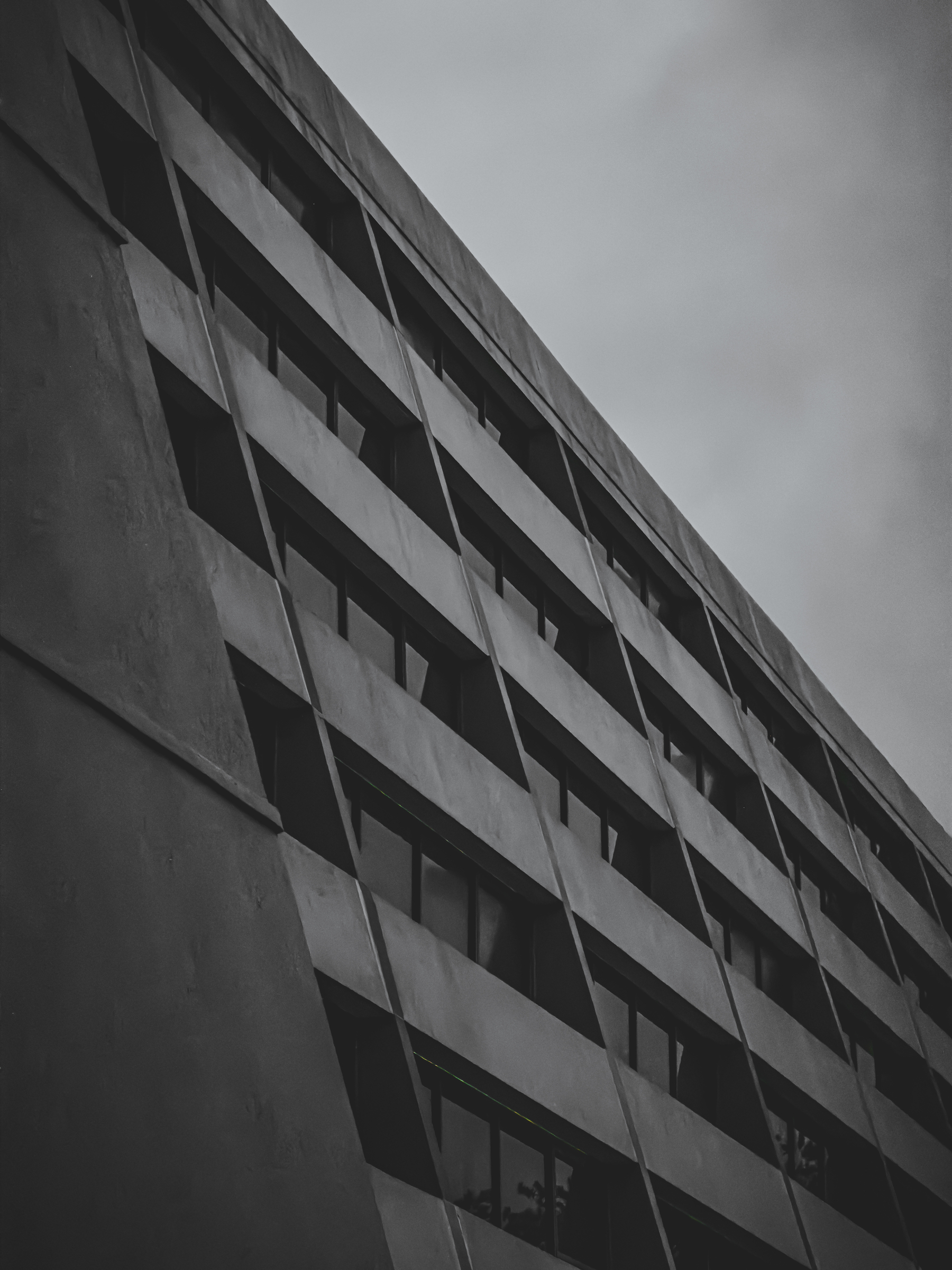 Auto-generated description: A grayscale photo shows the side of a modern multi-story building with rows of windows under a cloudy sky.