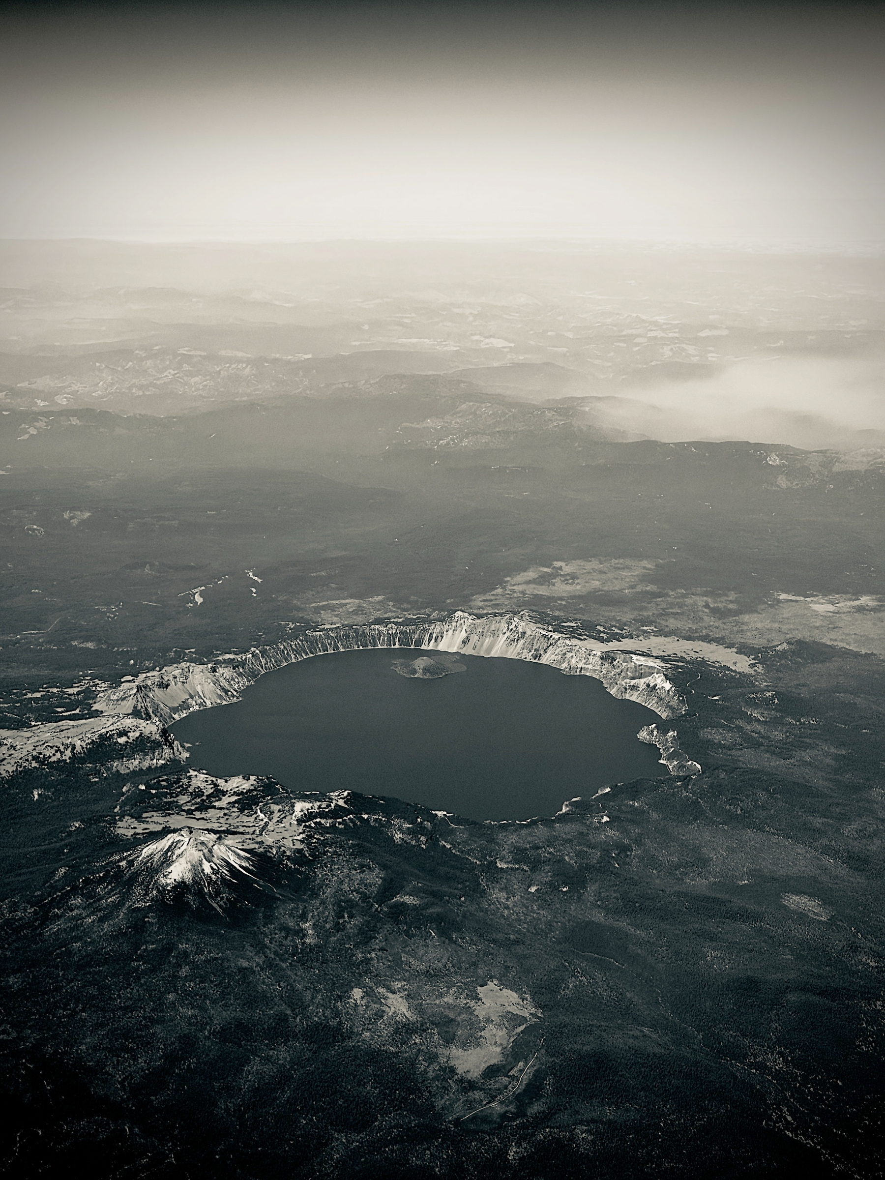 Auto-generated description: A large volcanic crater filled with water, surrounded by rugged mountainous terrain, is seen from an aerial perspective in a monochrome setting.