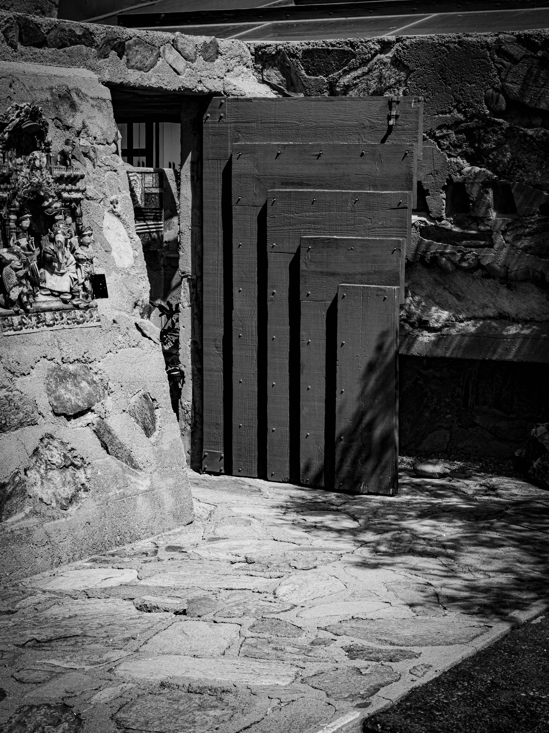 Auto-generated description: A large, layered geometric sculpture is positioned against a stone wall in a shadowed outdoor space, with a small religious shrine nearby.