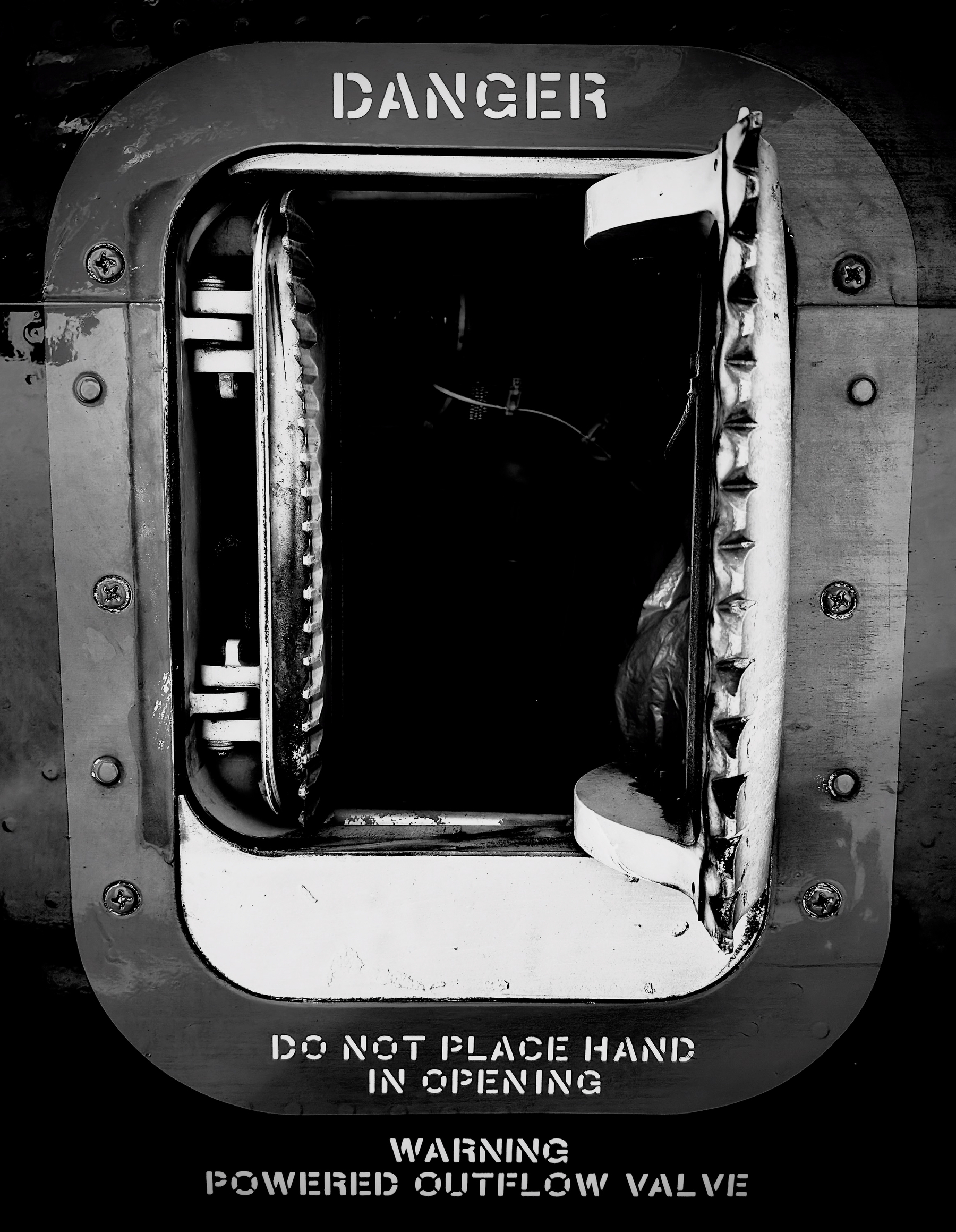 Auto-generated description: A metal hatch with warning labels indicating danger and caution against placing hands in the opening, featuring a partially open flap.