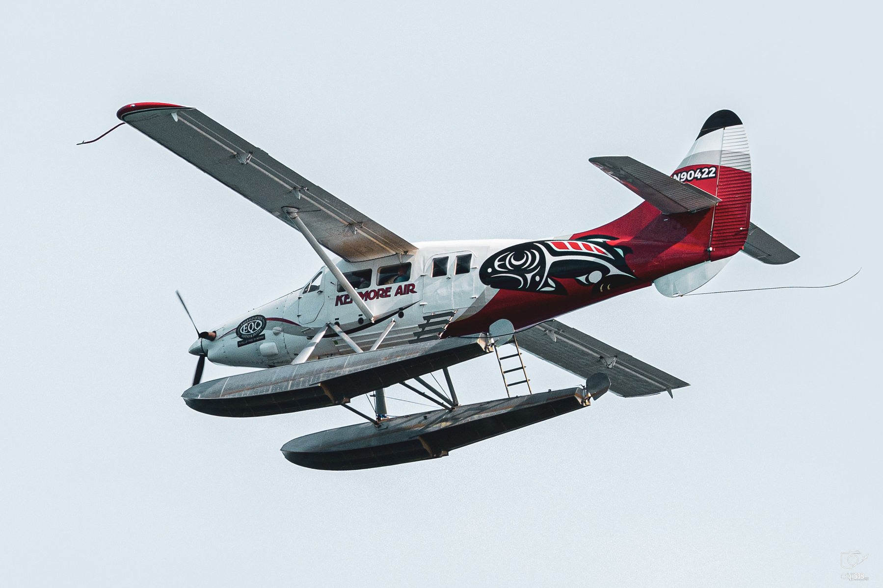 Auto-generated description: A floatplane with red, black, and white markings is flying against a clear sky.