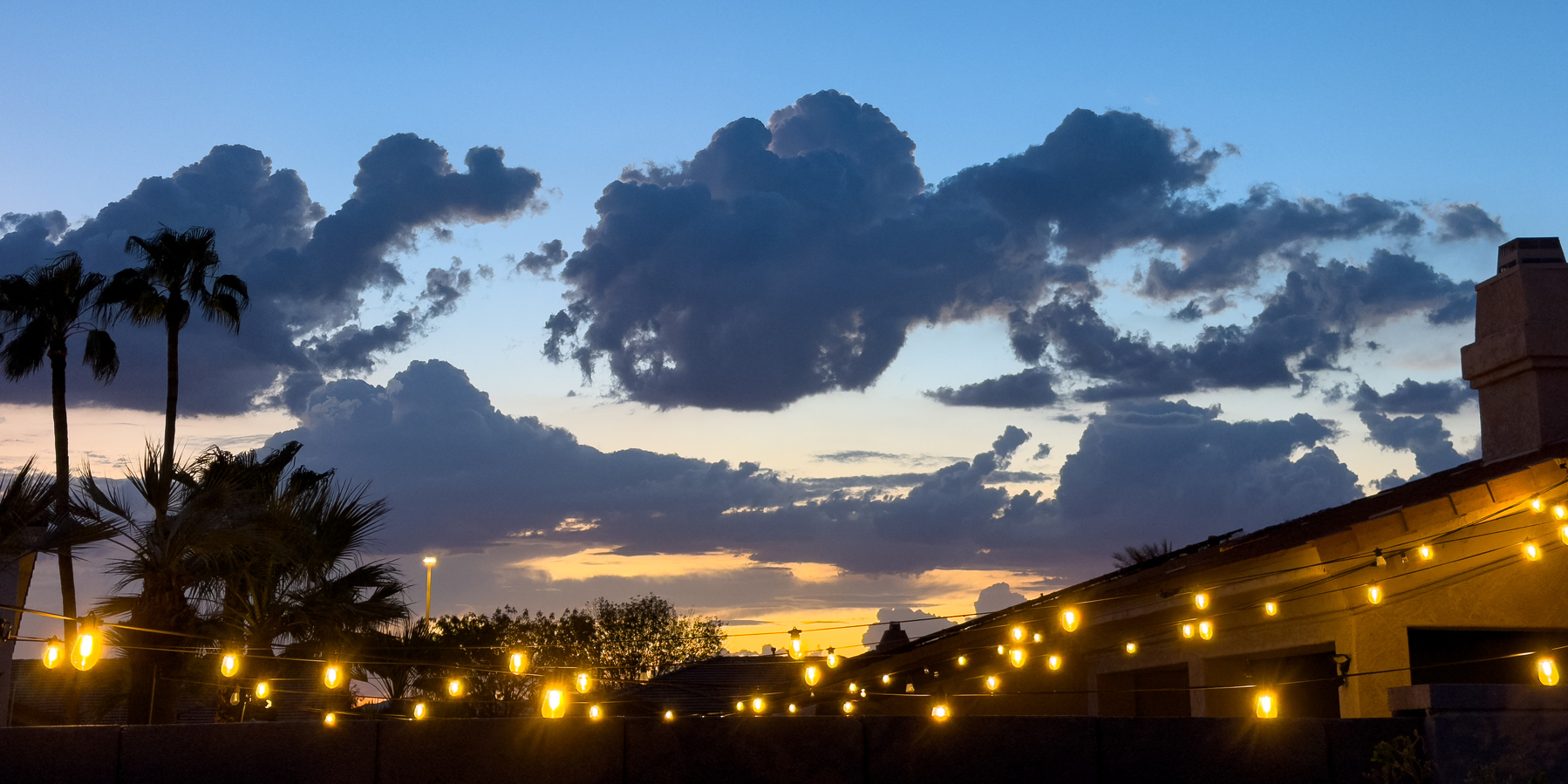 Auto-generated description: A sunset sky filled with dramatic clouds is seen above palm trees and string lights hanging along the roofline of a building.