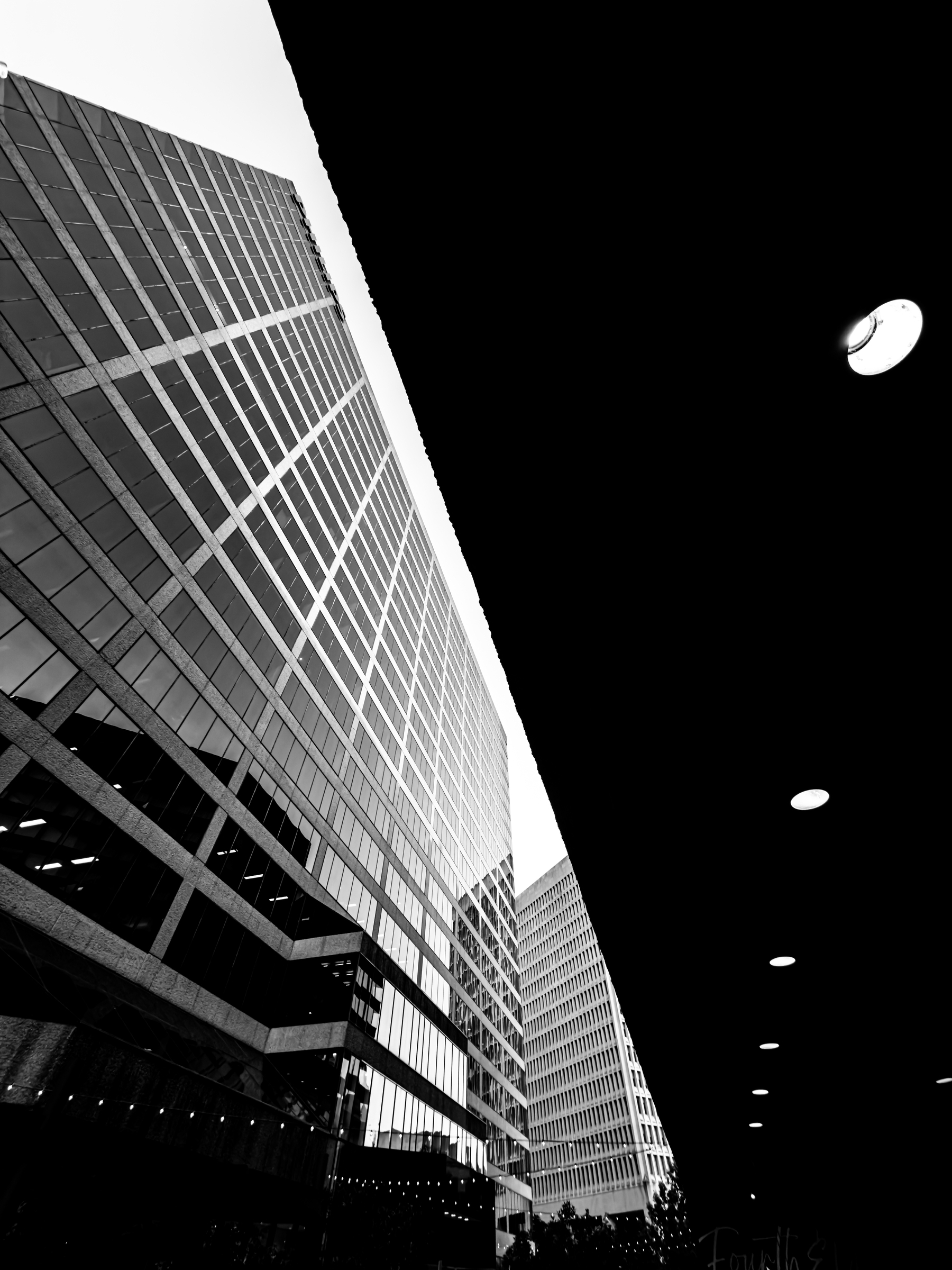 Auto-generated description: Tall modern office buildings are captured from a low angle with an overhang and ceiling lights framing the right side in a high-contrast black-and-white photo.