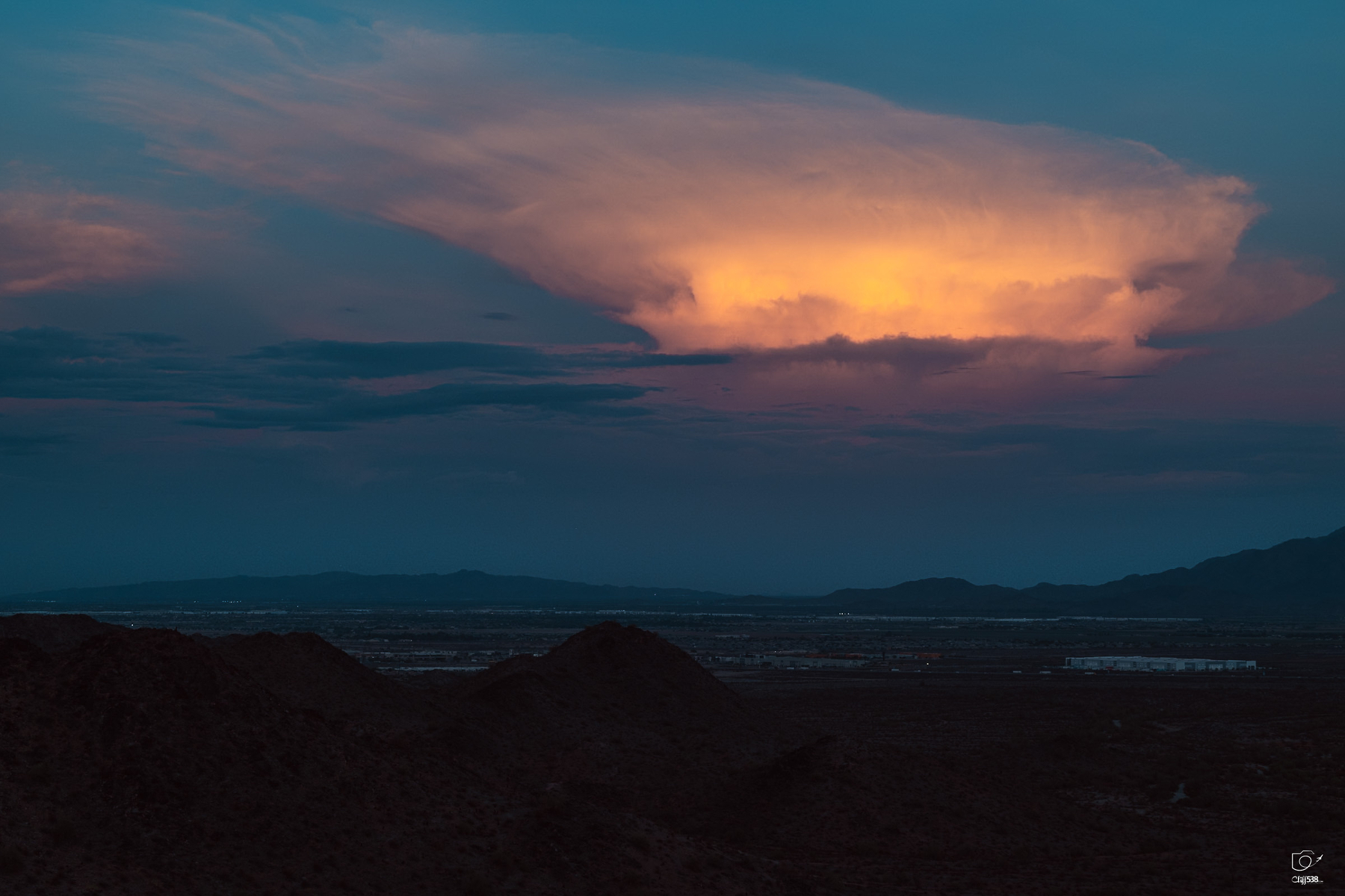 Auto-generated description: A dramatic sunset illuminates a large, towering cloud formation above a dark mountainous landscape.