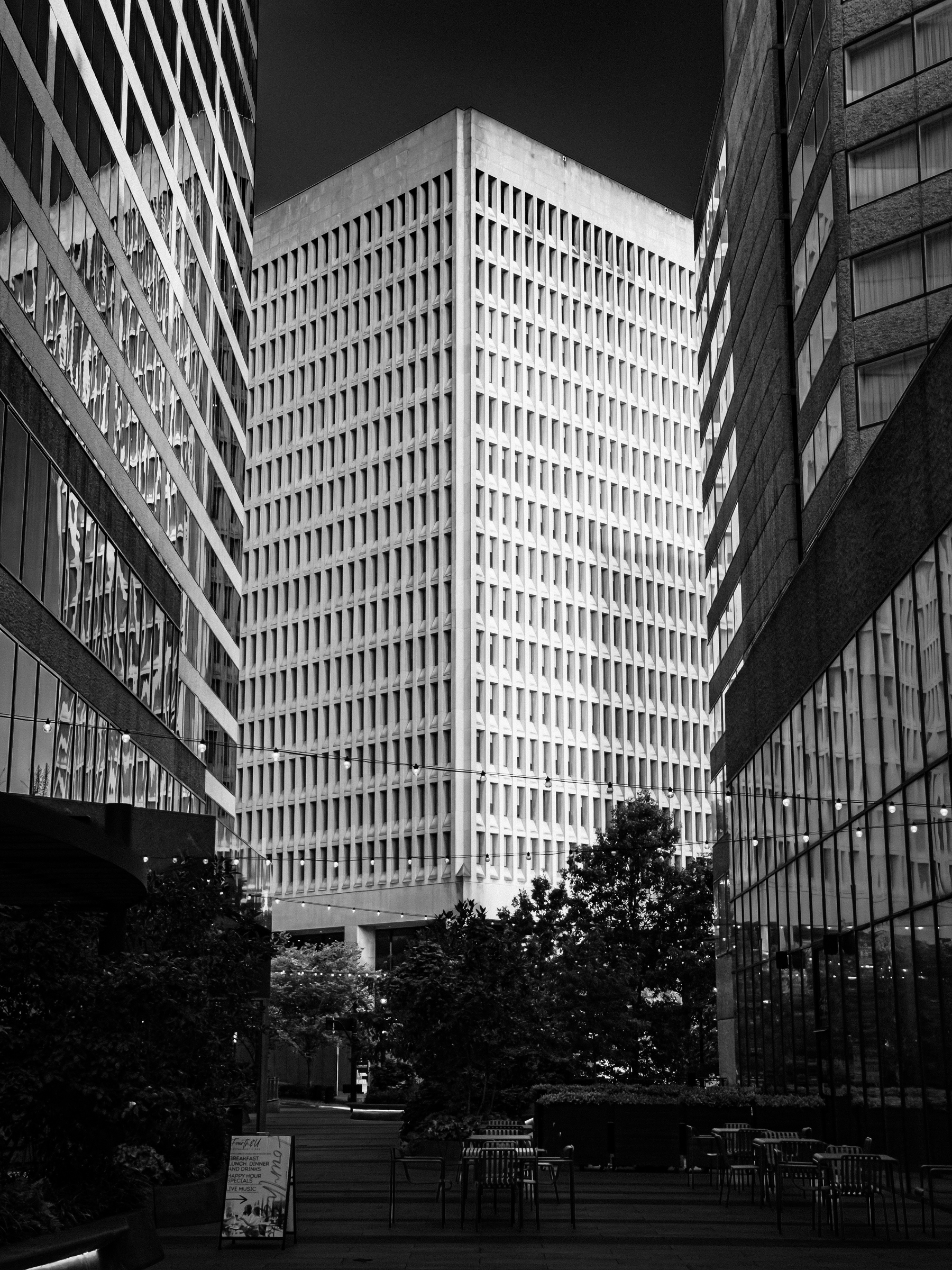 Auto-generated description: A tall, rectangular building with a grid-like window pattern is framed by two modern glass buildings on either side, with trees and outdoor seating visible in the foreground.