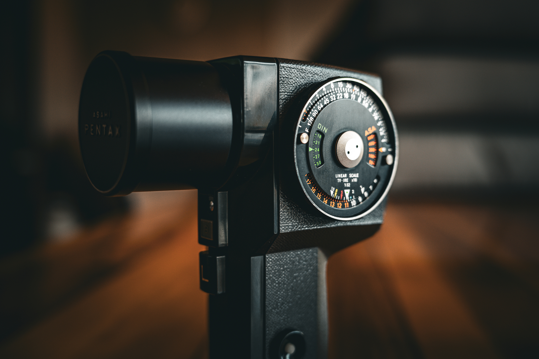 Auto-generated description: A vintage Pentax light meter is positioned on a wooden surface with a blurred background.