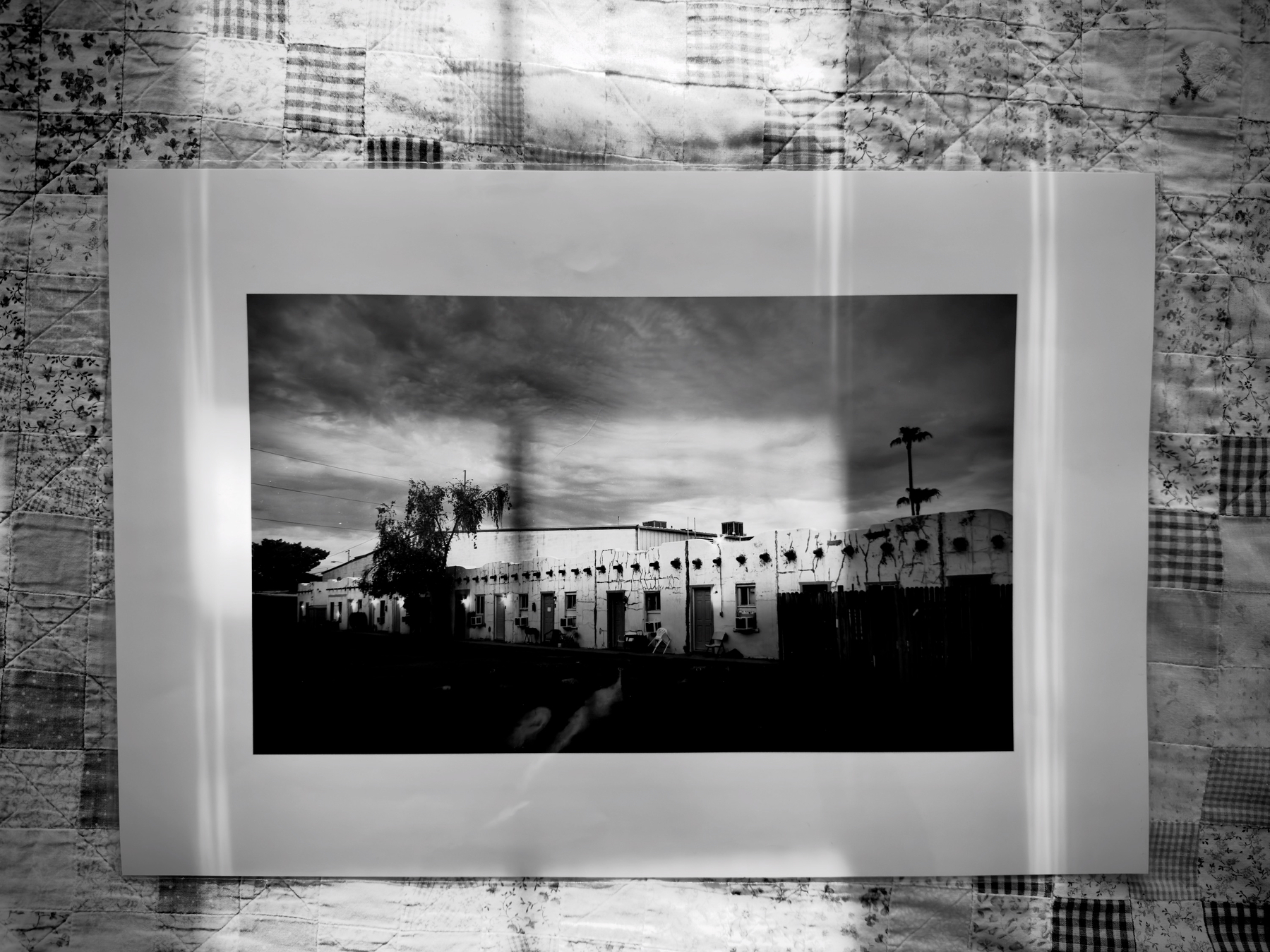 Auto-generated description: A black-and-white photograph of a building is mounted on a patterned quilted background with shadows cast across it.