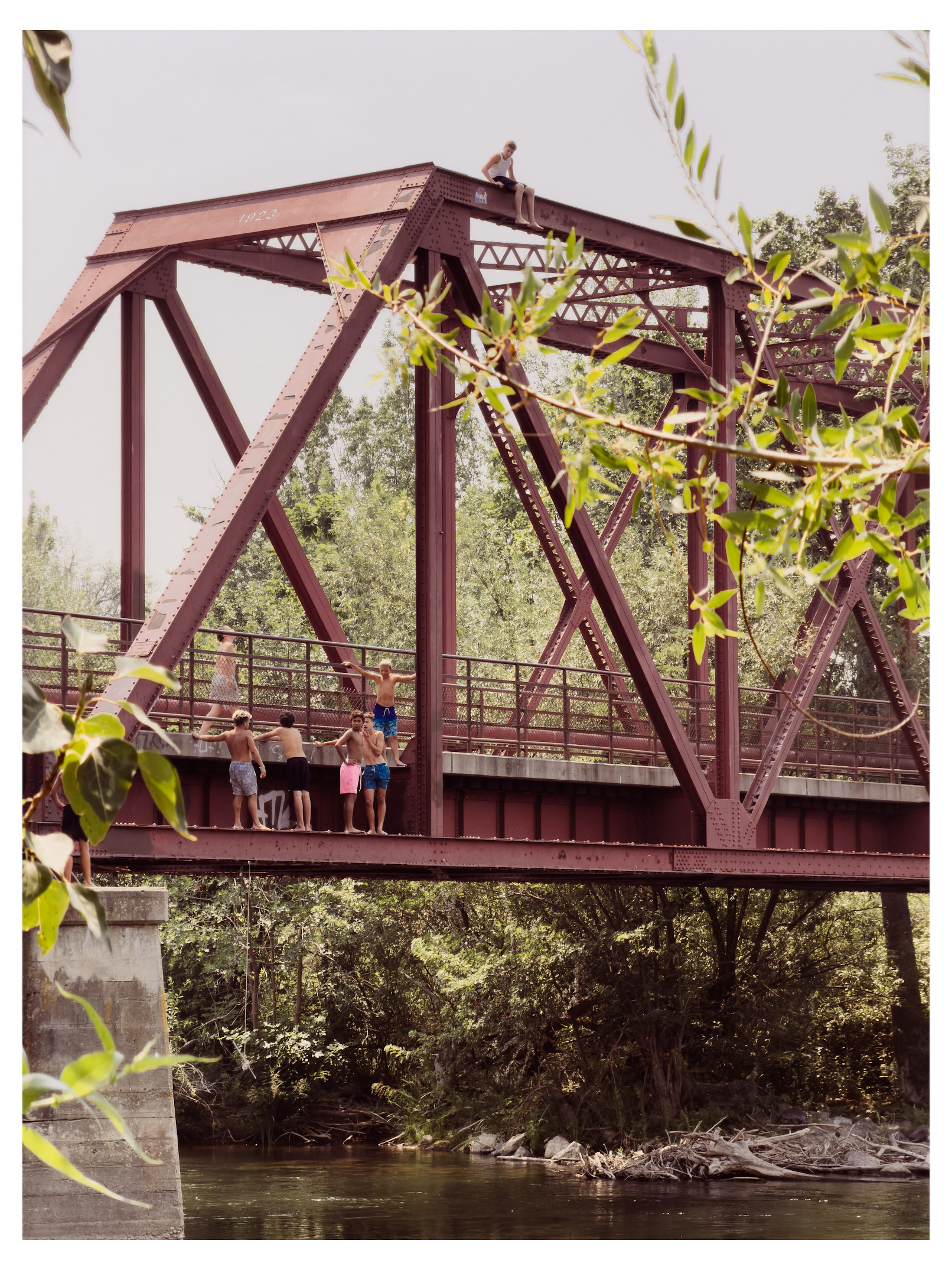Auto-generated description: A group of people, mostly children in swimwear, are gathered on and around a red metal bridge over a river, with one person sitting on top of the bridge structure.