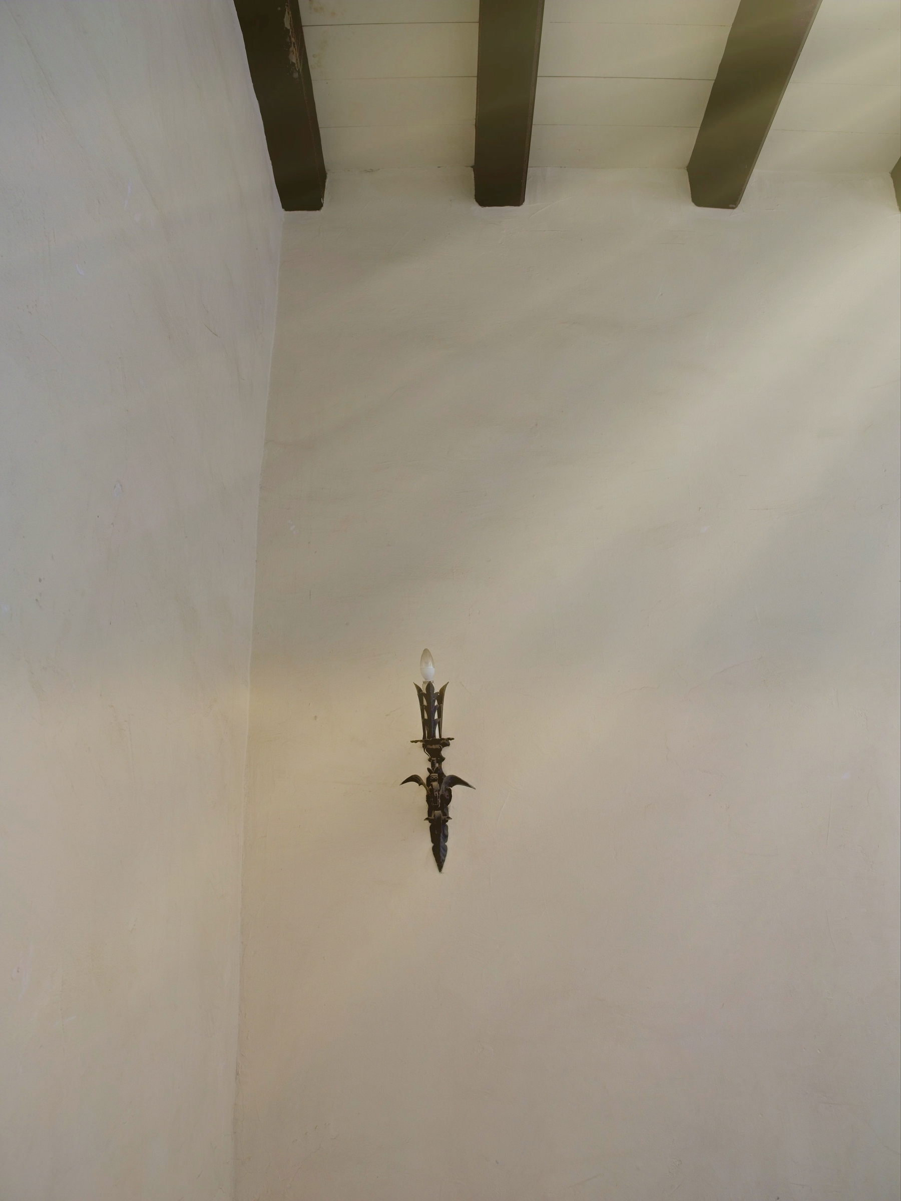 A decorative wall sconce with an unlit candle is mounted on a textured cream-colored wall beneath a ceiling with dark wooden beams.