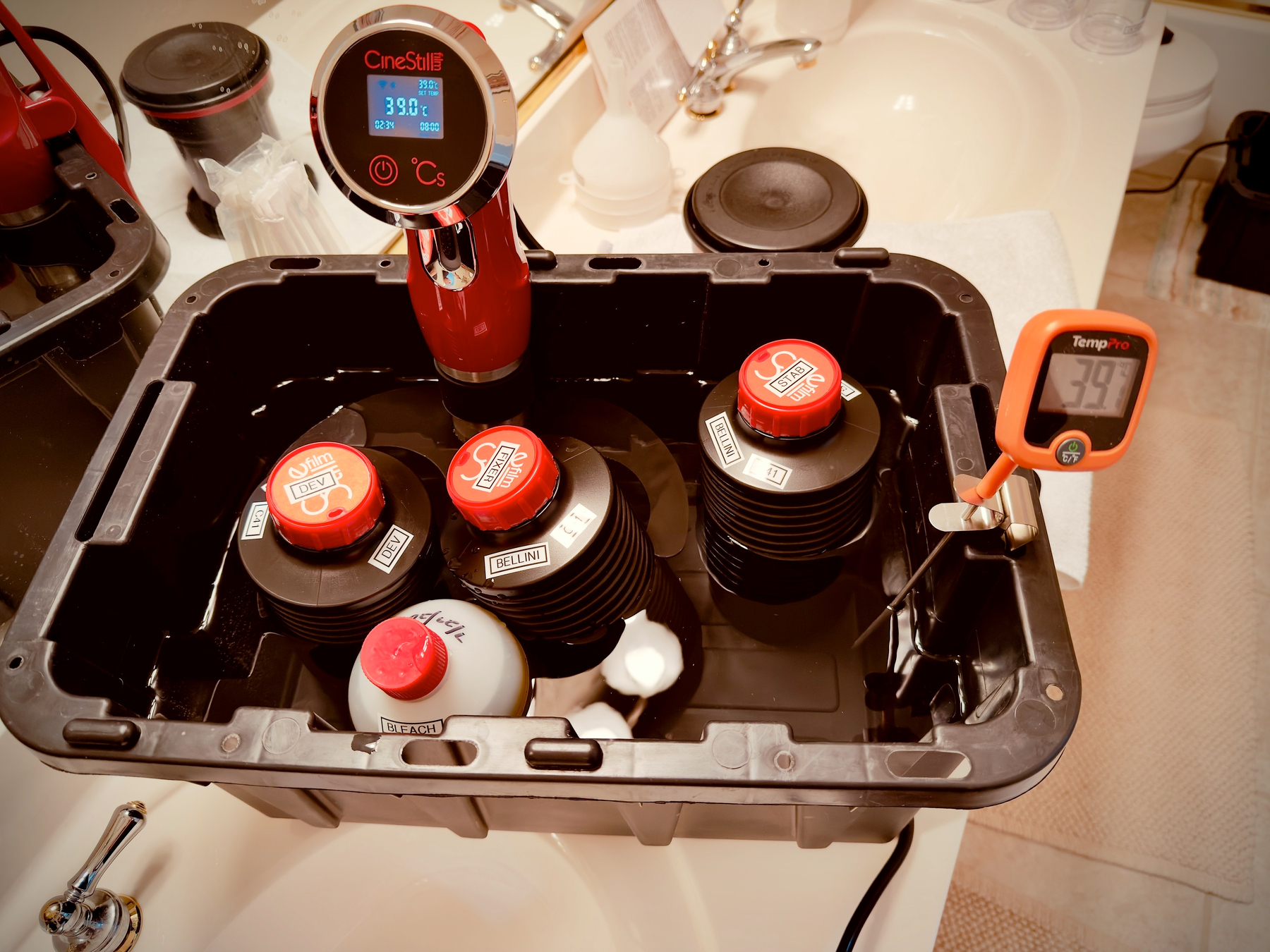 Auto-generated description: A film developing tank setup with multiple reels submerged in liquid, featuring a digital temperature meter and a thermometer attached to the container.