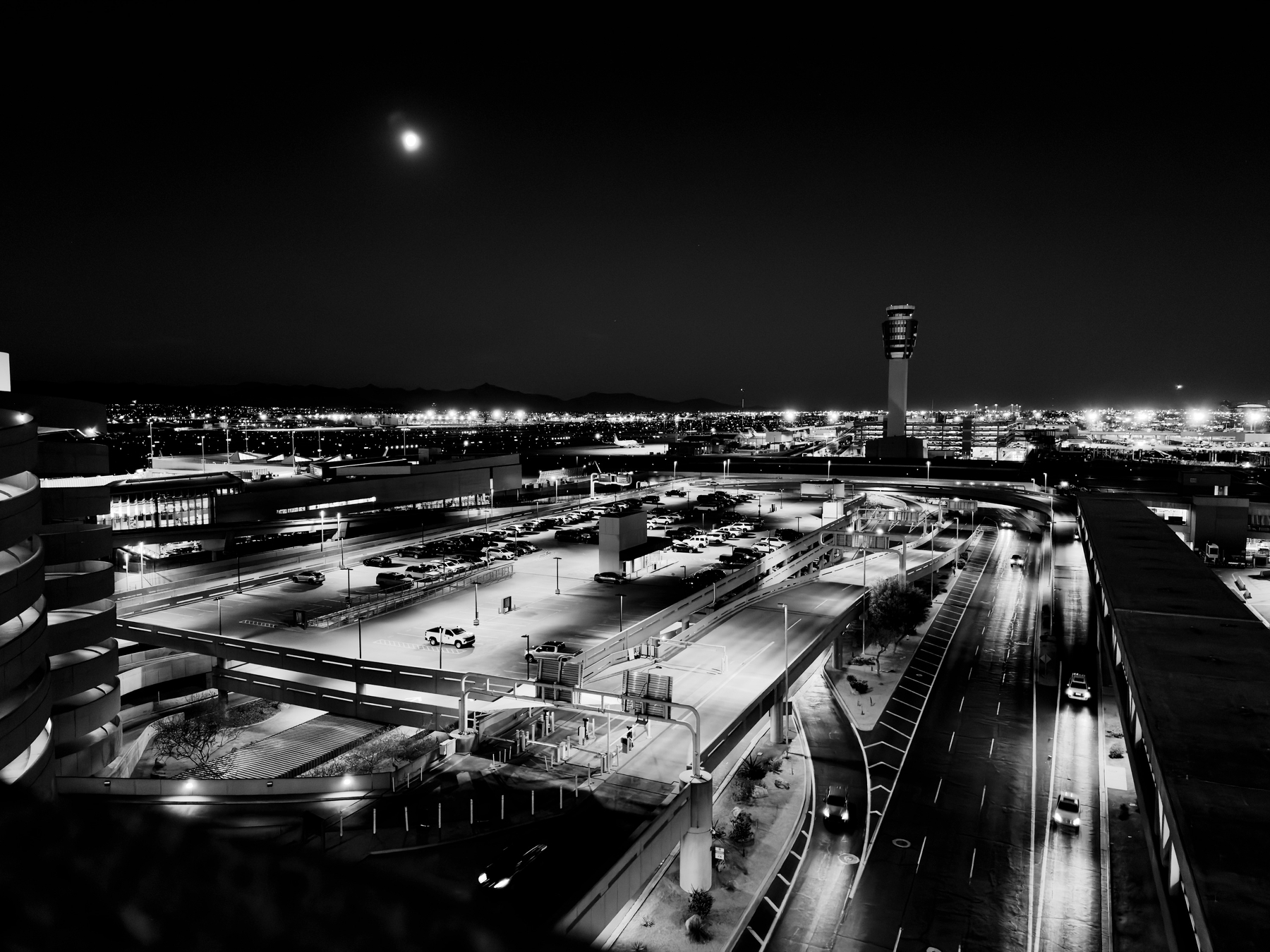 Auto-generated description: A nighttime aerial view of an airport with illuminated runways, control tower, and surrounding city lights beneath a moonlit sky.