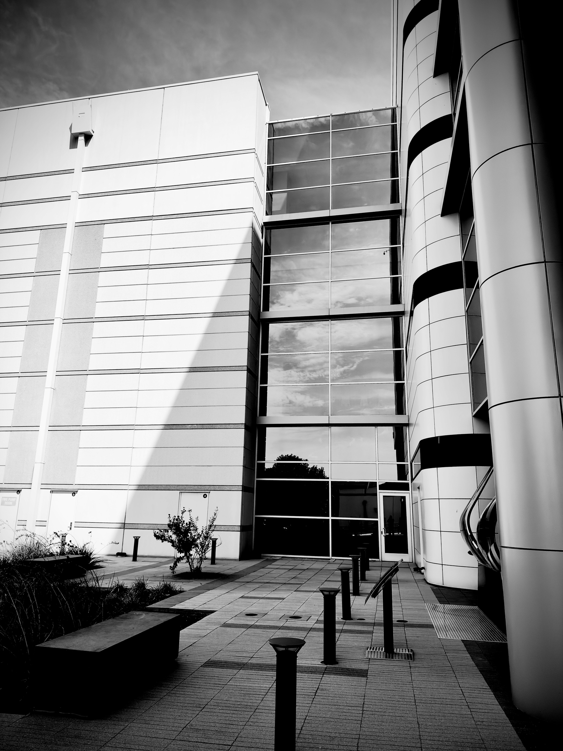 Auto-generated description: A modern building exterior features large glass windows, cylindrical structural elements, and a paved walkway with several bollards and a bench.