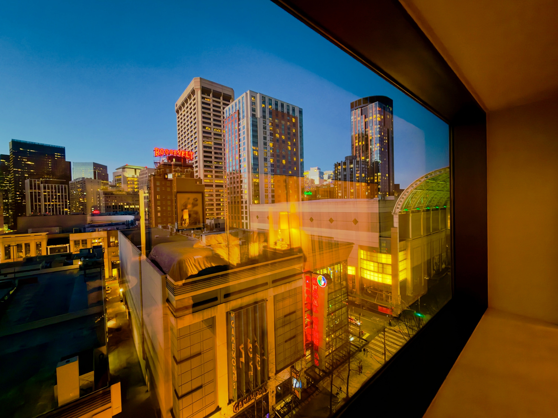 Looking through a window, a vibrant cityscape with illuminated skyscrapers and buildings under a twilight sky is visible.