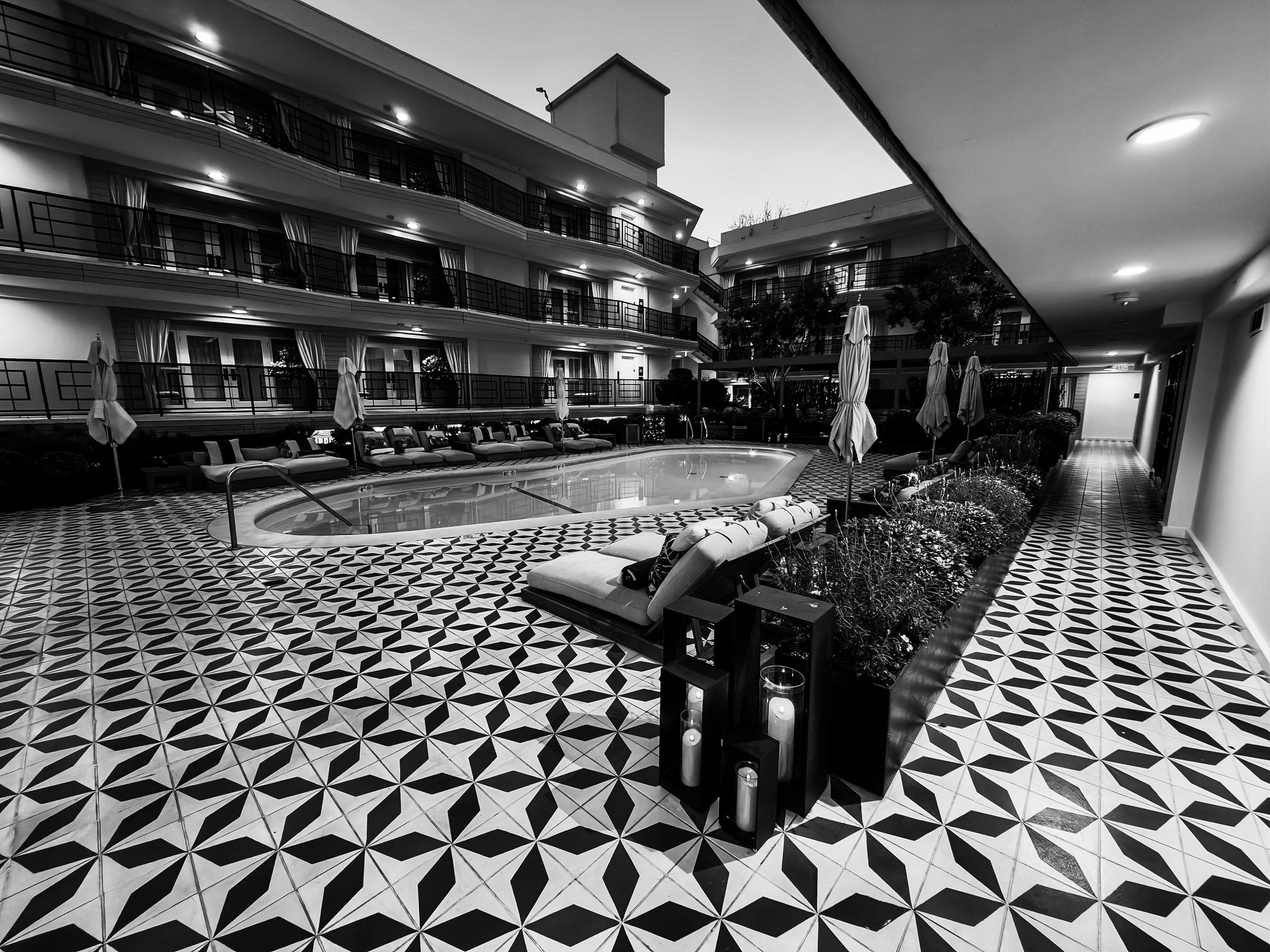 A black and white photograph depicts a modern hotel courtyard with a geometric-tiled floor, a swimming pool, and several poolside chairs and umbrellas.