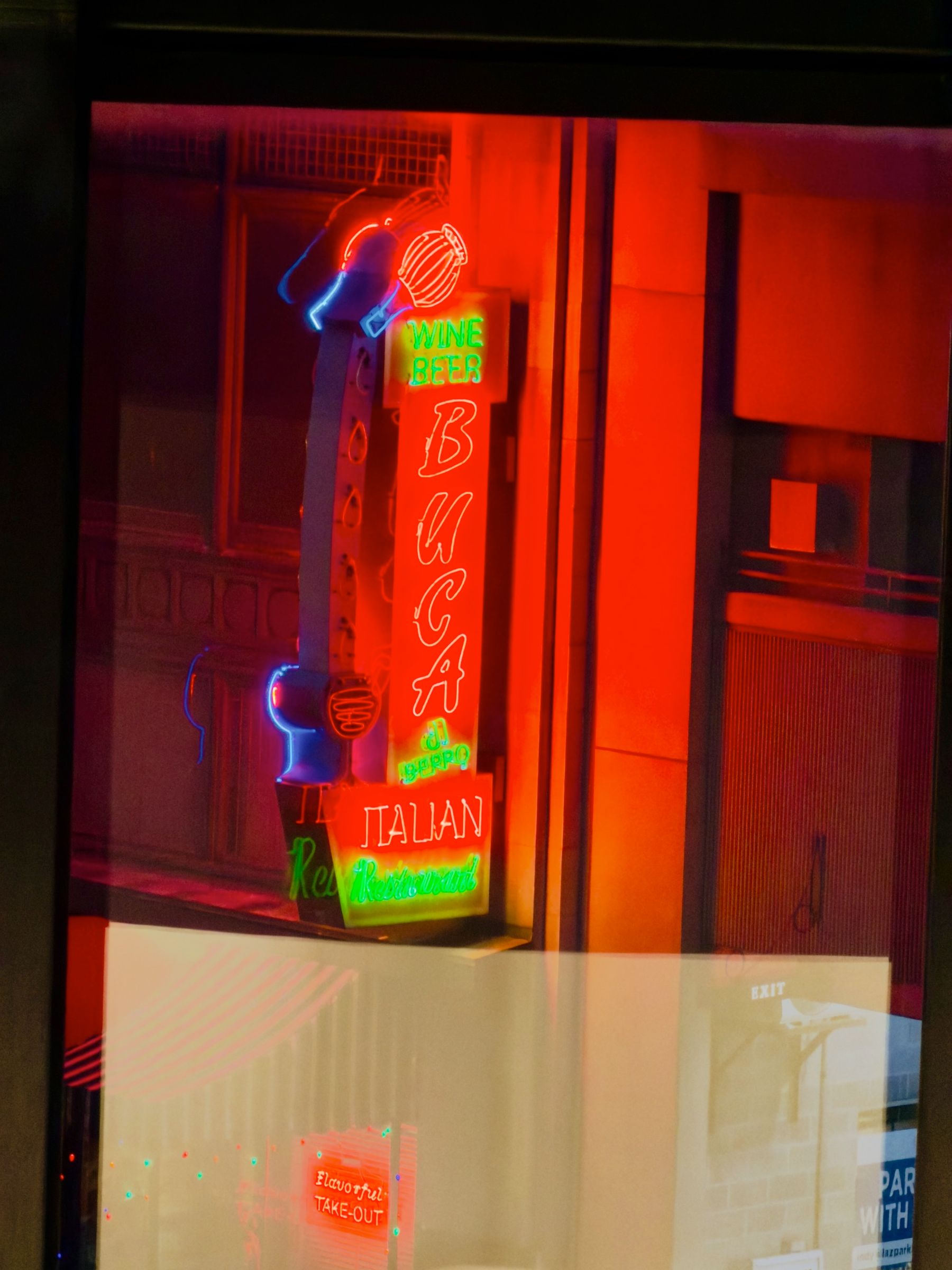 Auto-generated description: A neon sign advertising Buca, an Italian restaurant with wine, glows vibrantly in shades of red, blue, and green against an urban backdrop.