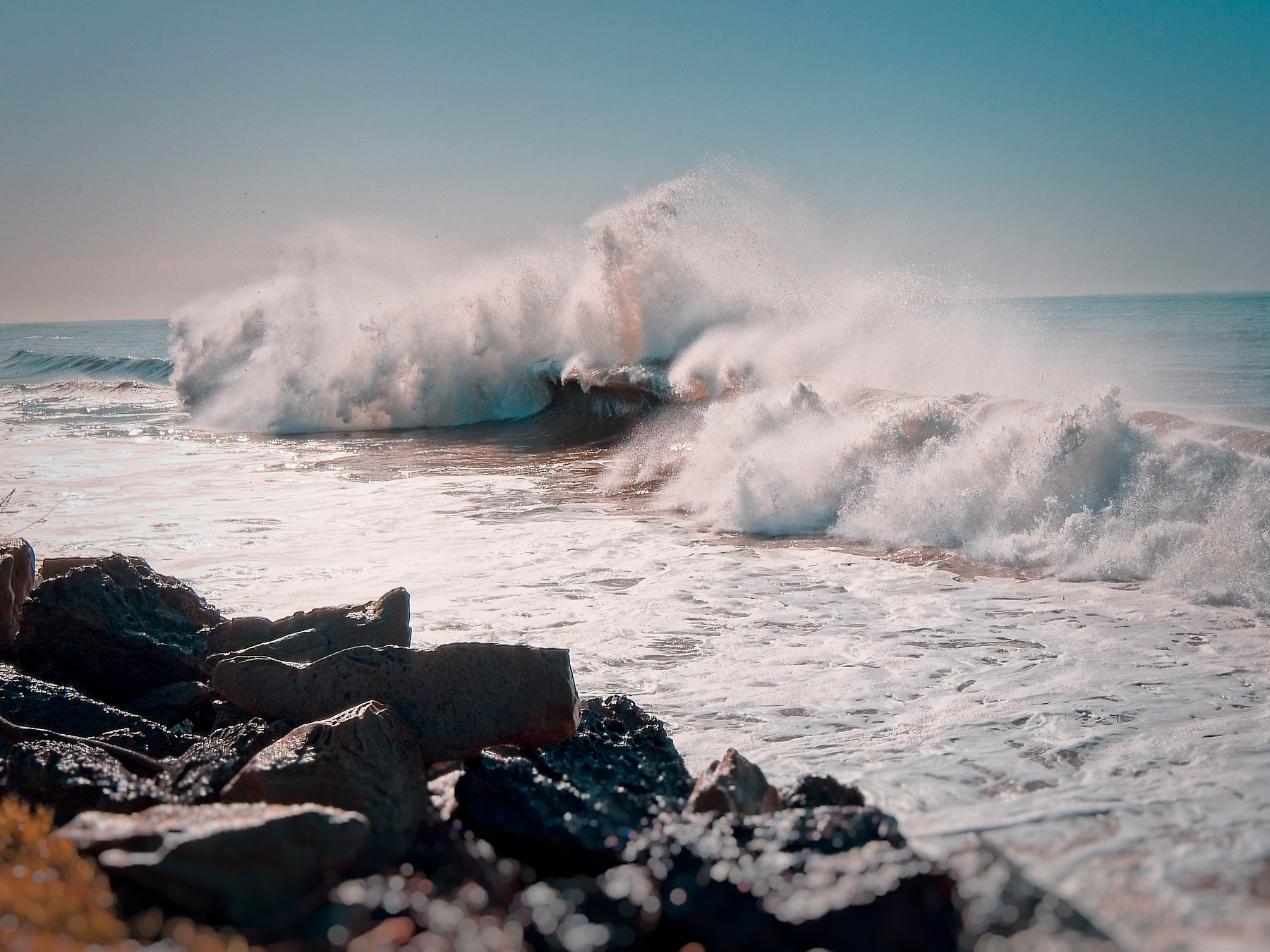 Powerful ocean waves crash against rocky shores under a clear sky.