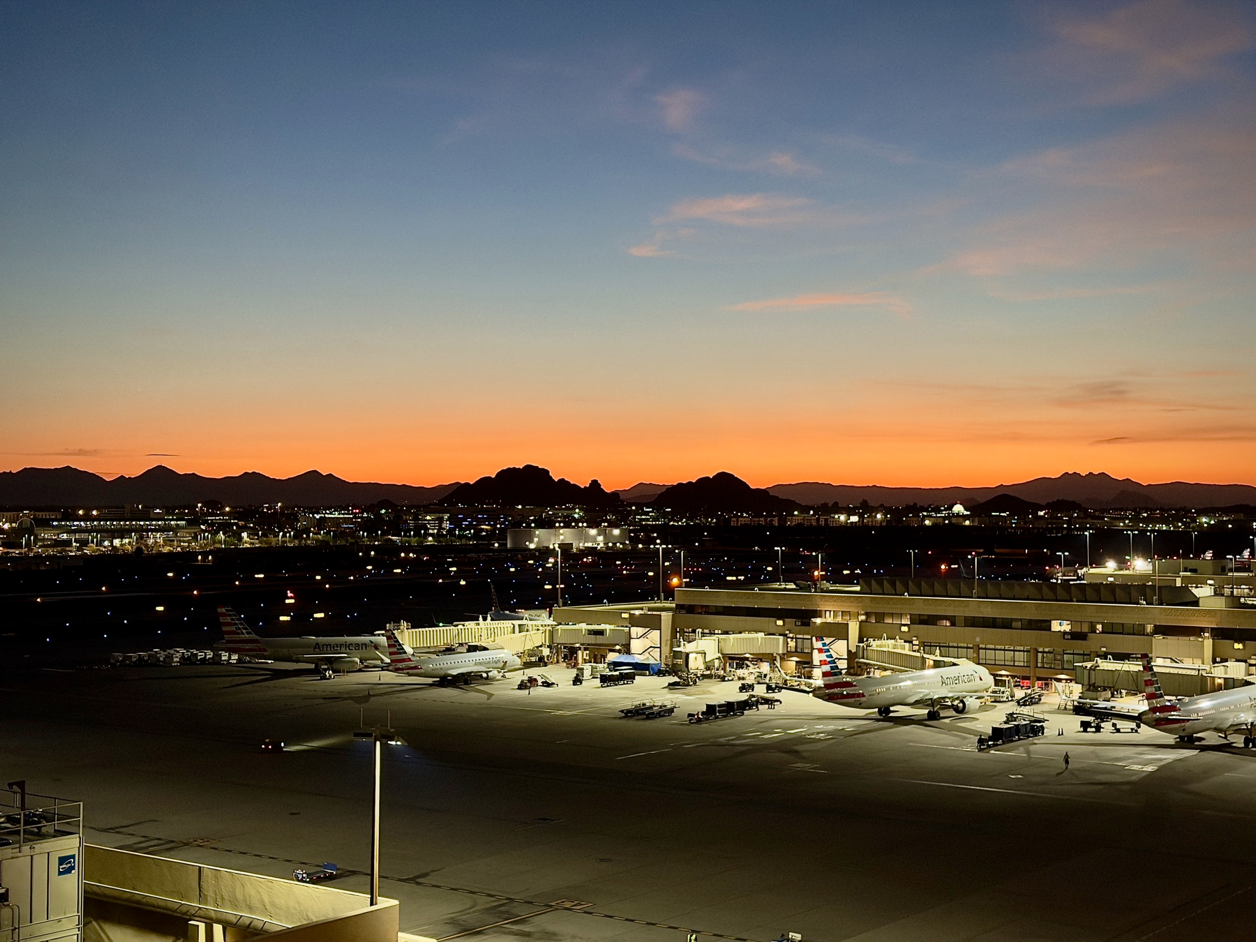 Auto-generated description: An airport with several airplanes parked at gates during sunset with mountains visible in the background.