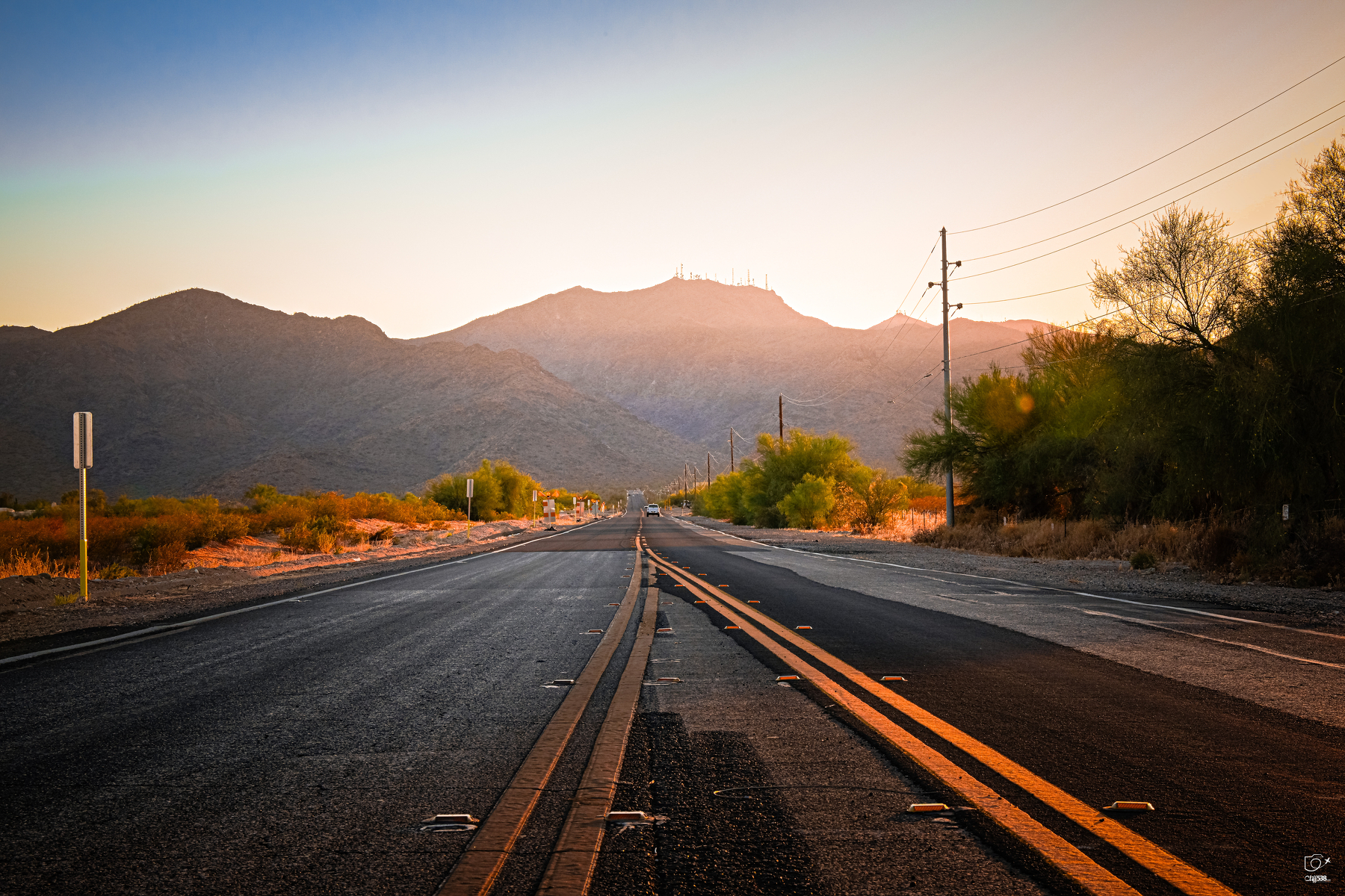 Auto-generated description: A long, empty road stretches toward a mountain range under a clear sky during sunset.