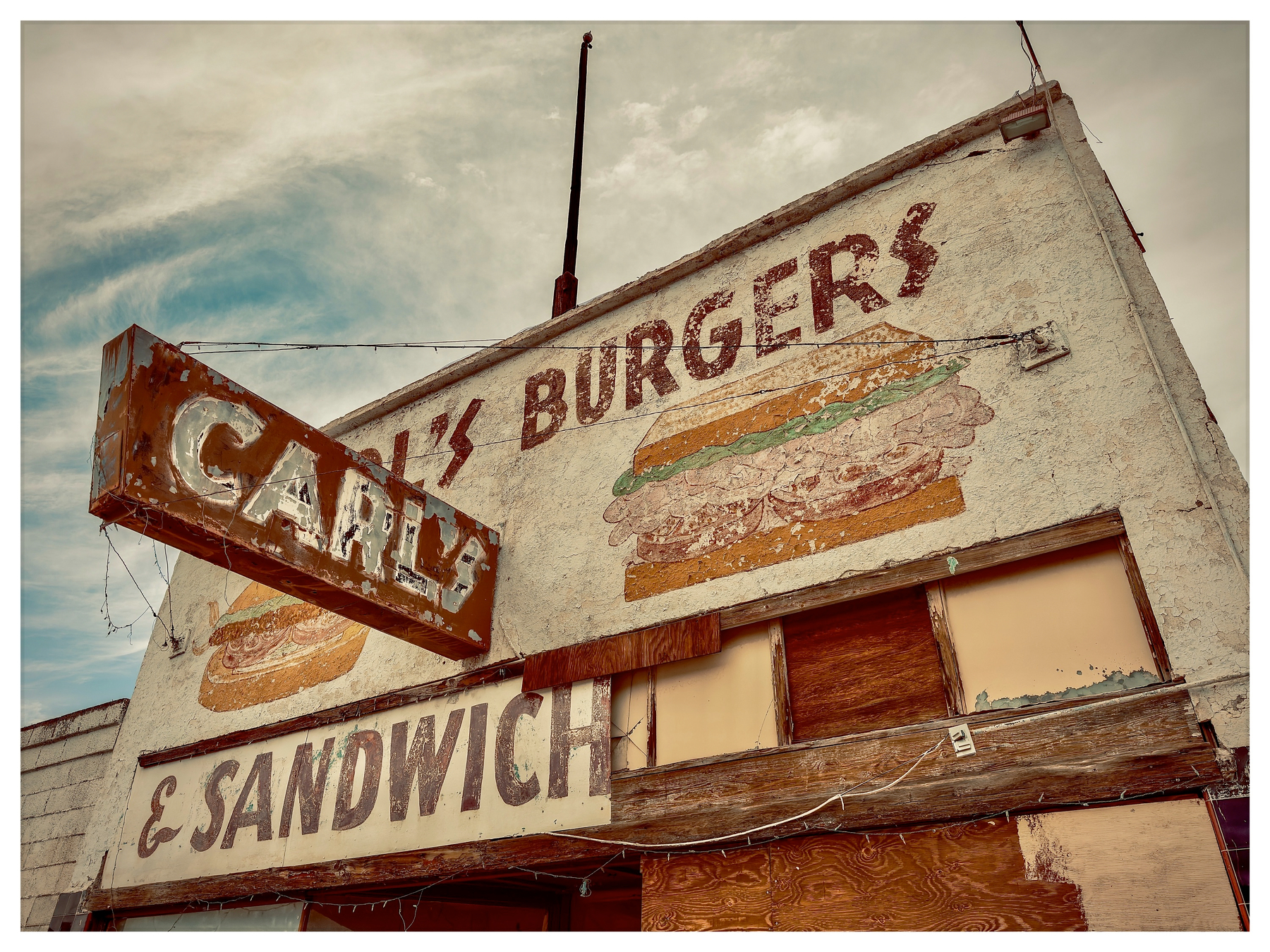 Auto-generated description: A weathered and partially boarded-up building displays faded signage for Carl's Burgers & Sandwiches with illustrations of burgers.