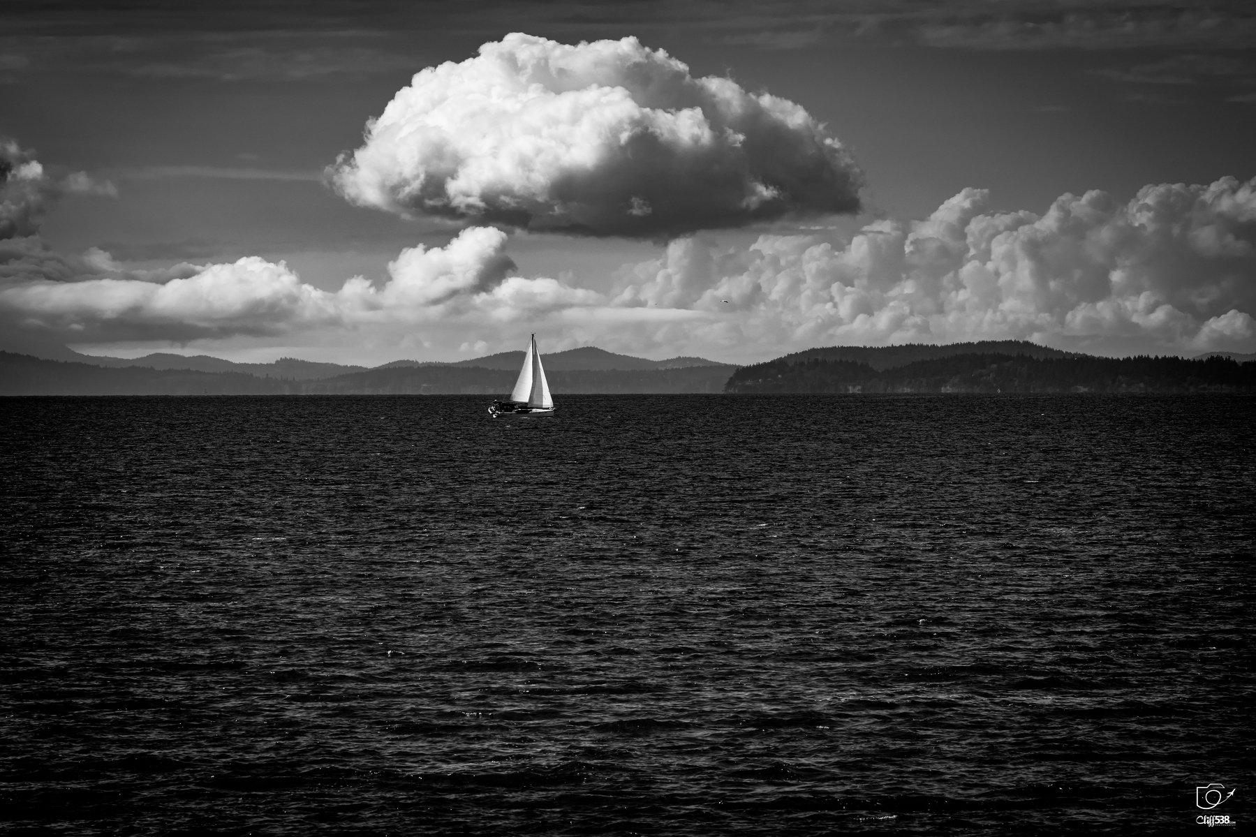 Auto-generated description: A lone sailboat glides across calm waters with a dramatic cloudy sky and distant landforms in the background.