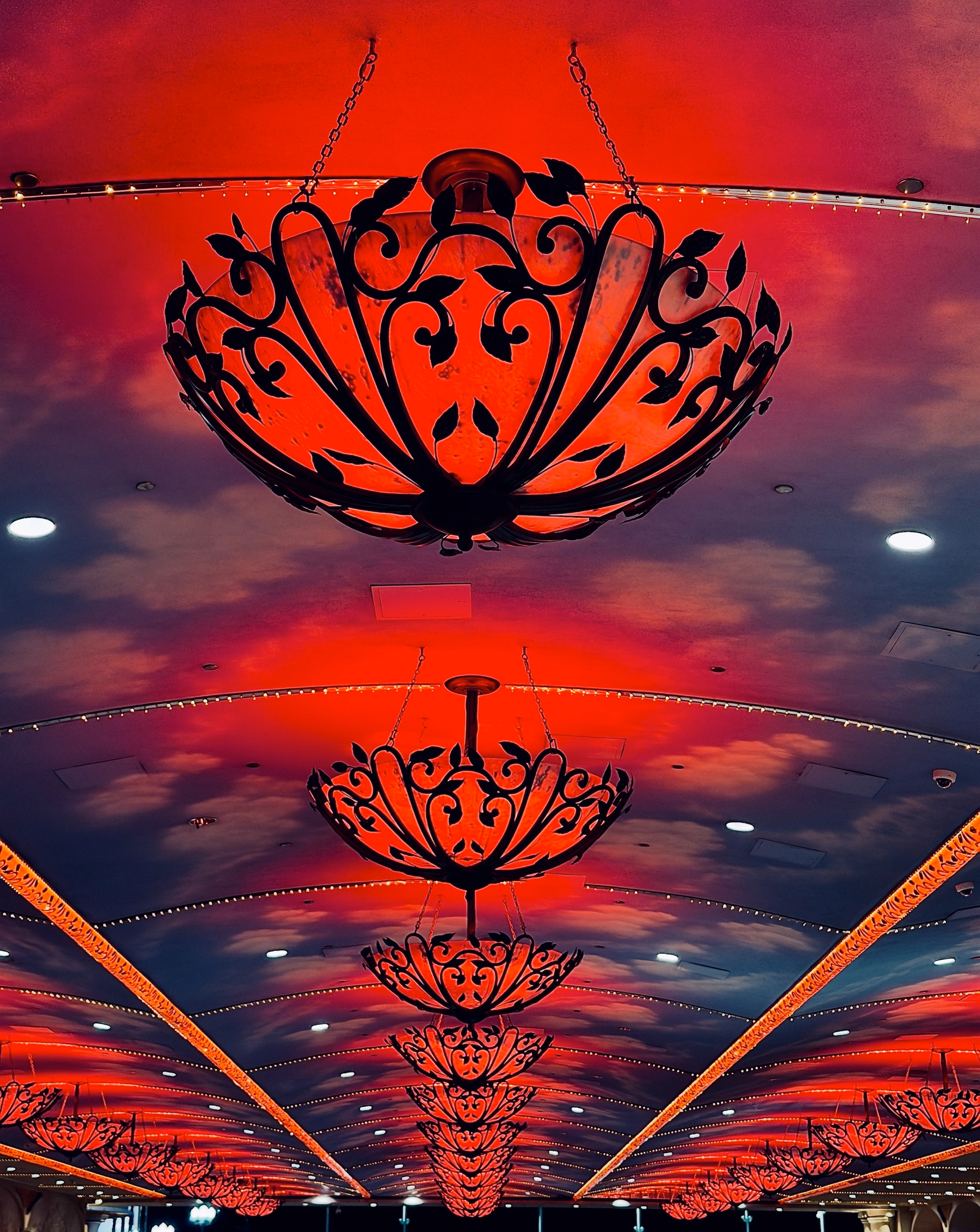 Auto-generated description: A series of ornate chandeliers with intricate black metal designs hang from a ceiling illuminated by warm red and orange lighting.