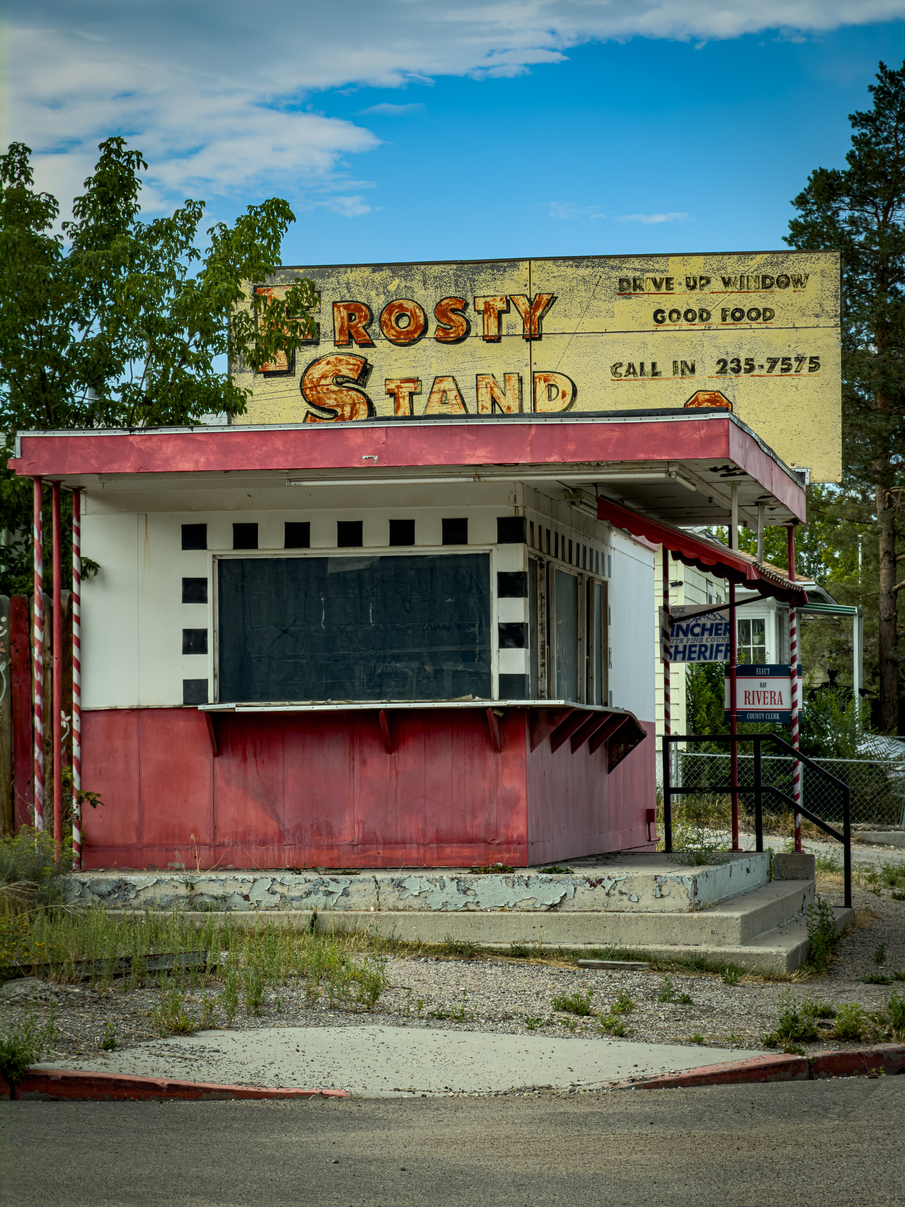 Auto-generated description: A small, weathered food stand named Frosty Stand with a faded sign advertising drive-up window service and a phone number sits abandoned in a grassy area.