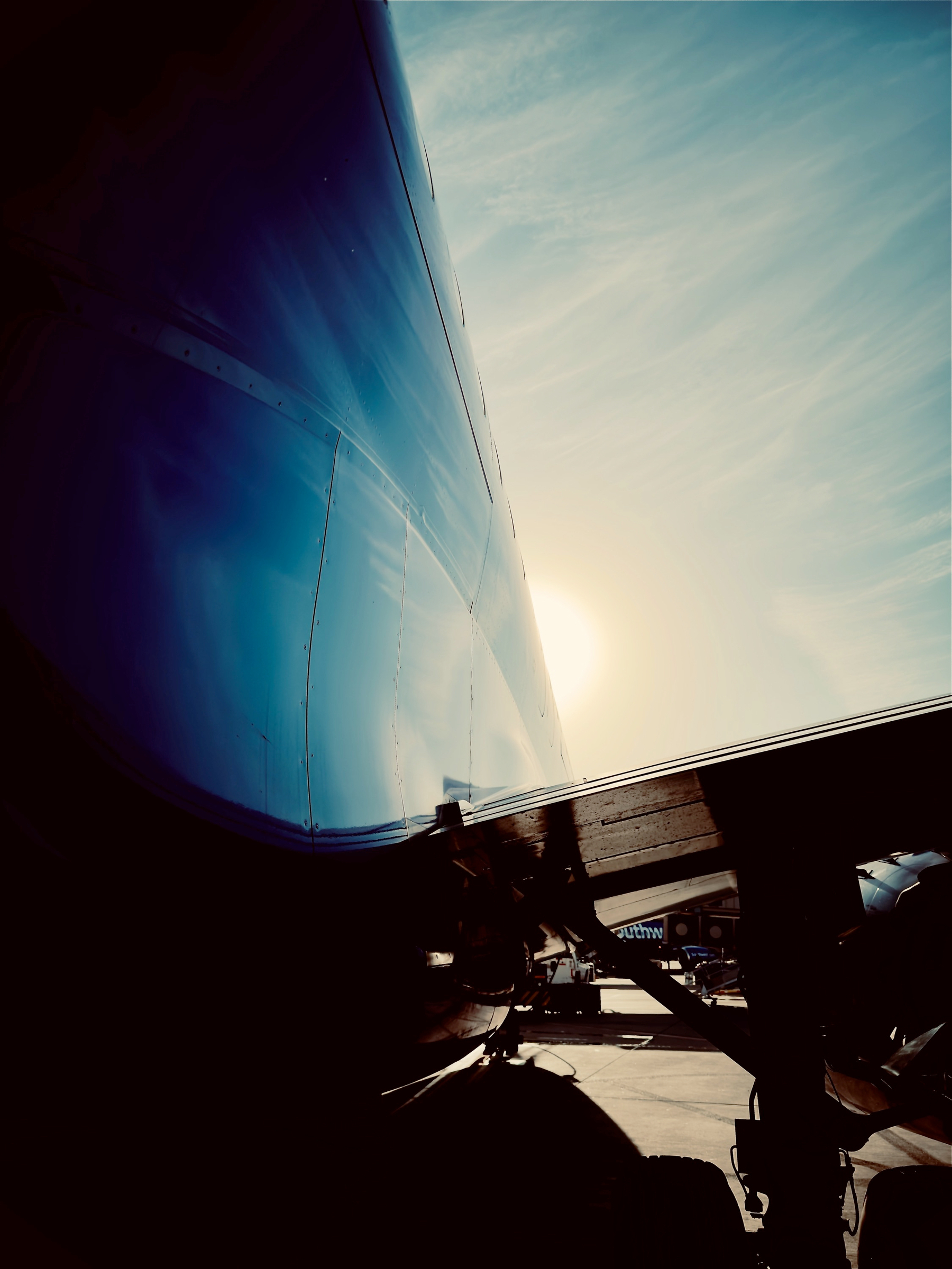 Auto-generated description: A close-up view of an airplane's body and landing gear is captured with the sun shining brightly in the background against a partly cloudy sky.